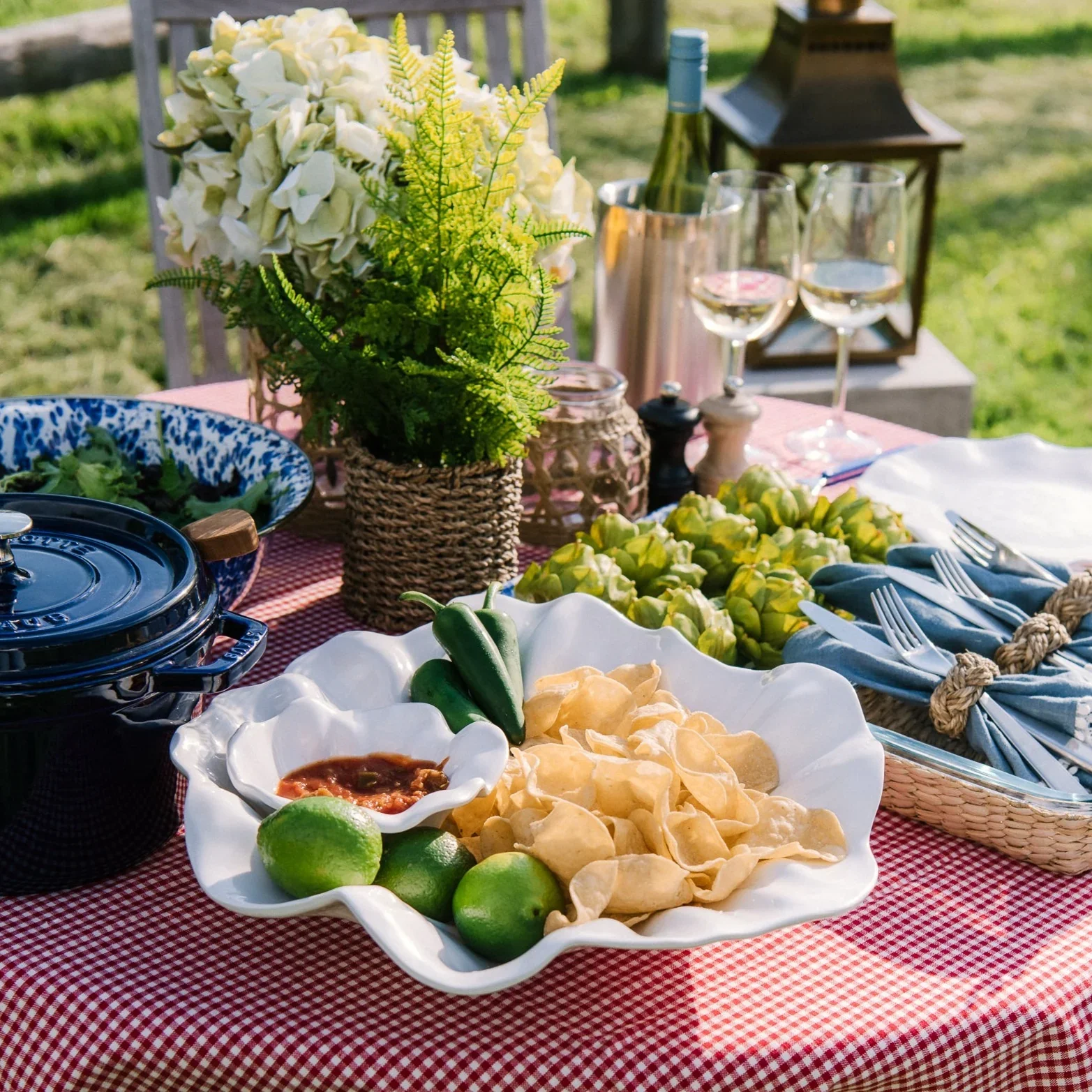 Outdoor table set with tortilla chips, guacamole, salsa, fresh limes, wine glasses, and a lantern.