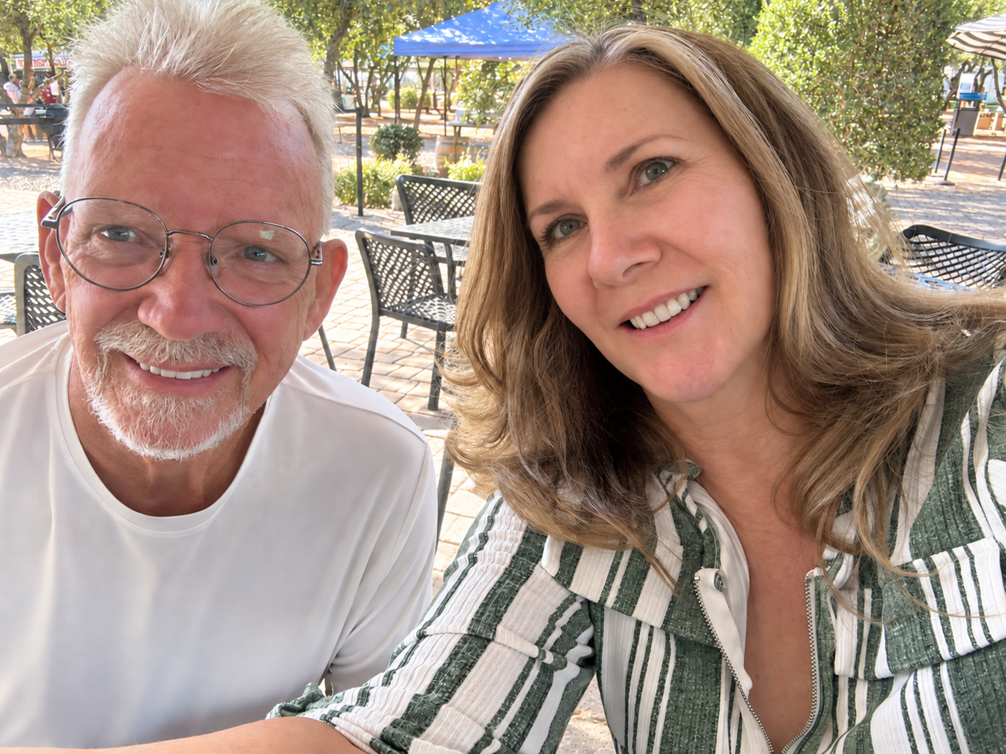 Karen and Johnny B smiling at an outdoor table beneath the olive trees at Queen Creek Olive Mill.