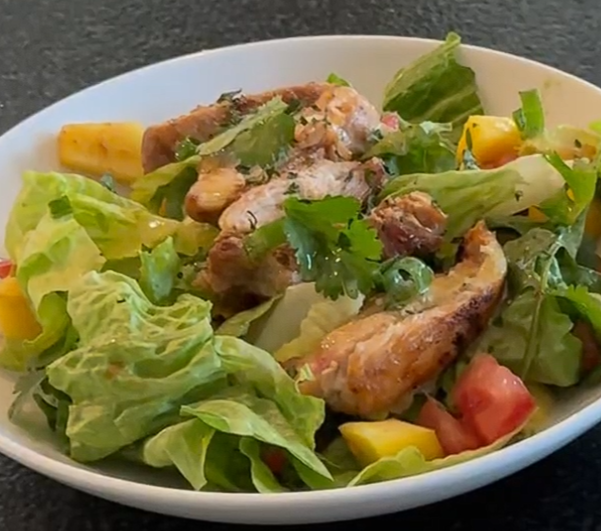 Mango chicken salad with grilled chicken, fresh mango, avocado, tomato, mixed greens, scallions, and cilantro in a white bowl.