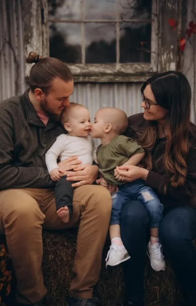 A man and a woman sitting together with two small children, touching noses, in a rustic setting with weathered wood and a window in the background.