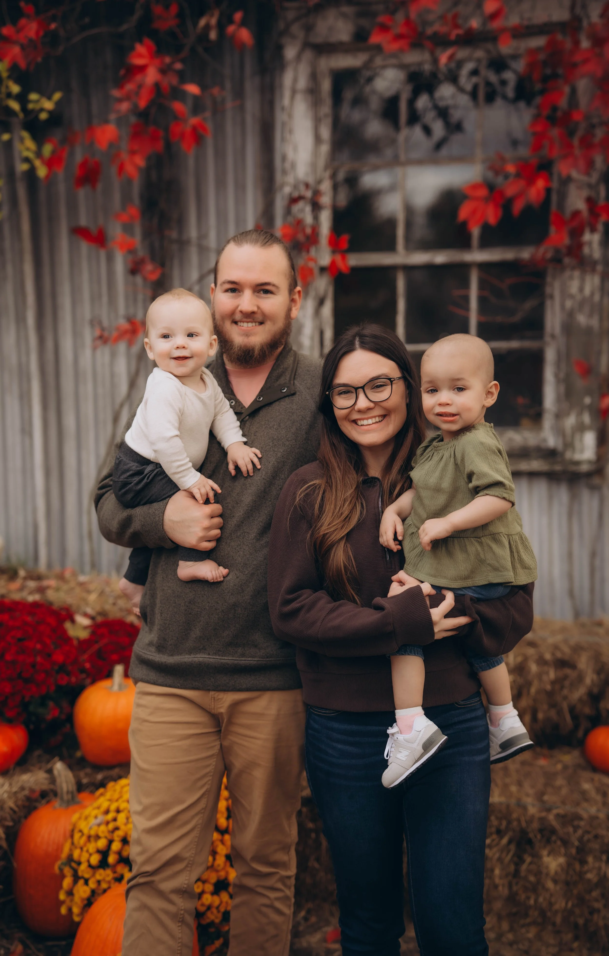 A smiling family of four outdoors with fall pumpkins and flowers, an old barn with red leaves behind them.