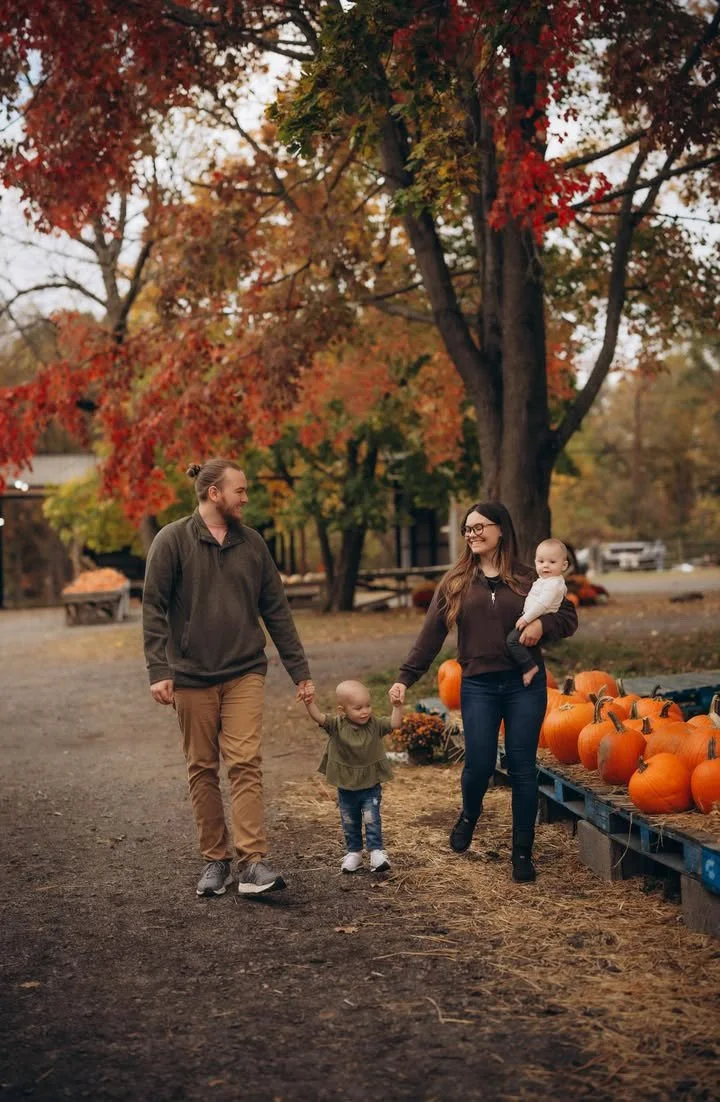 A family of four, including a man, woman, and two small children, walks hand-in-hand through a pumpkin patch with autumn trees and pumpkins in the background.