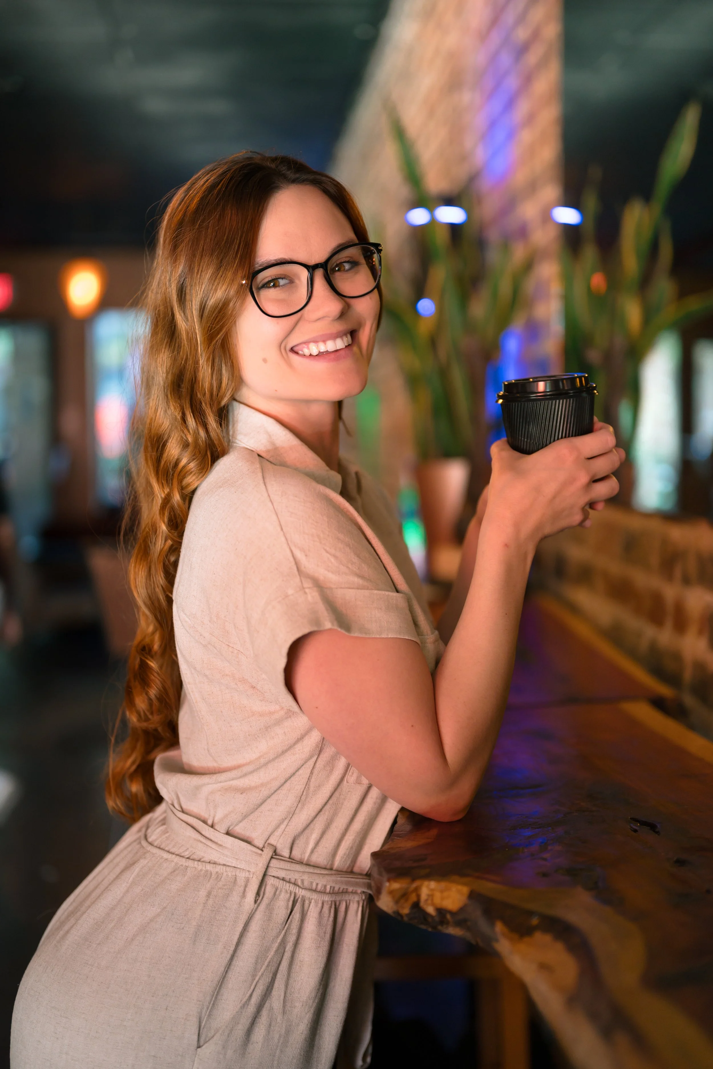 A woman with long red hair, glasses, and a beige outfit smiling while holding a black coffee cup in a cozy cafe with potted plants and brick walls.