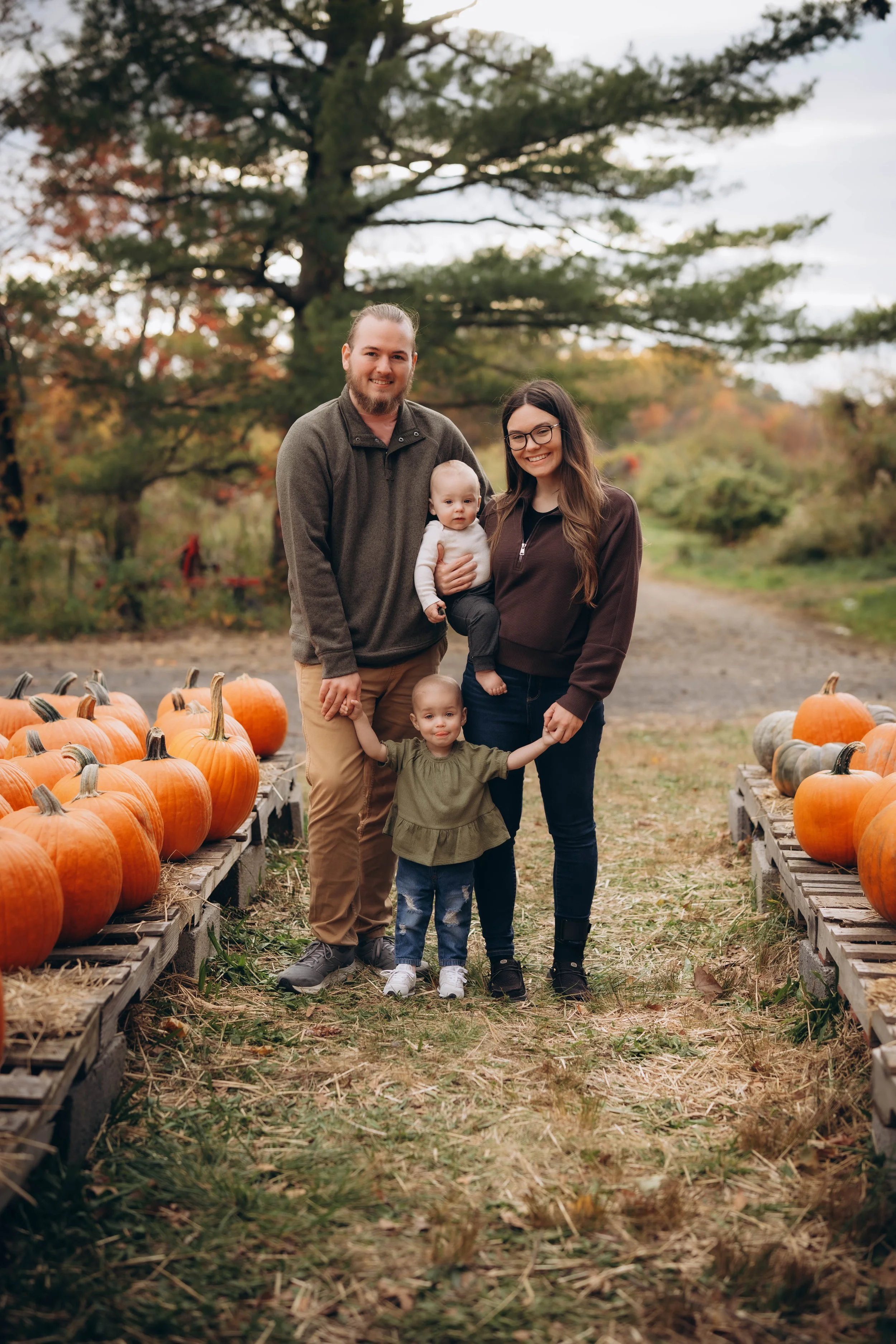 Family of four standing on a pumpkin patch path surrounded by pumpkins, with trees in autumn foliage in the background.