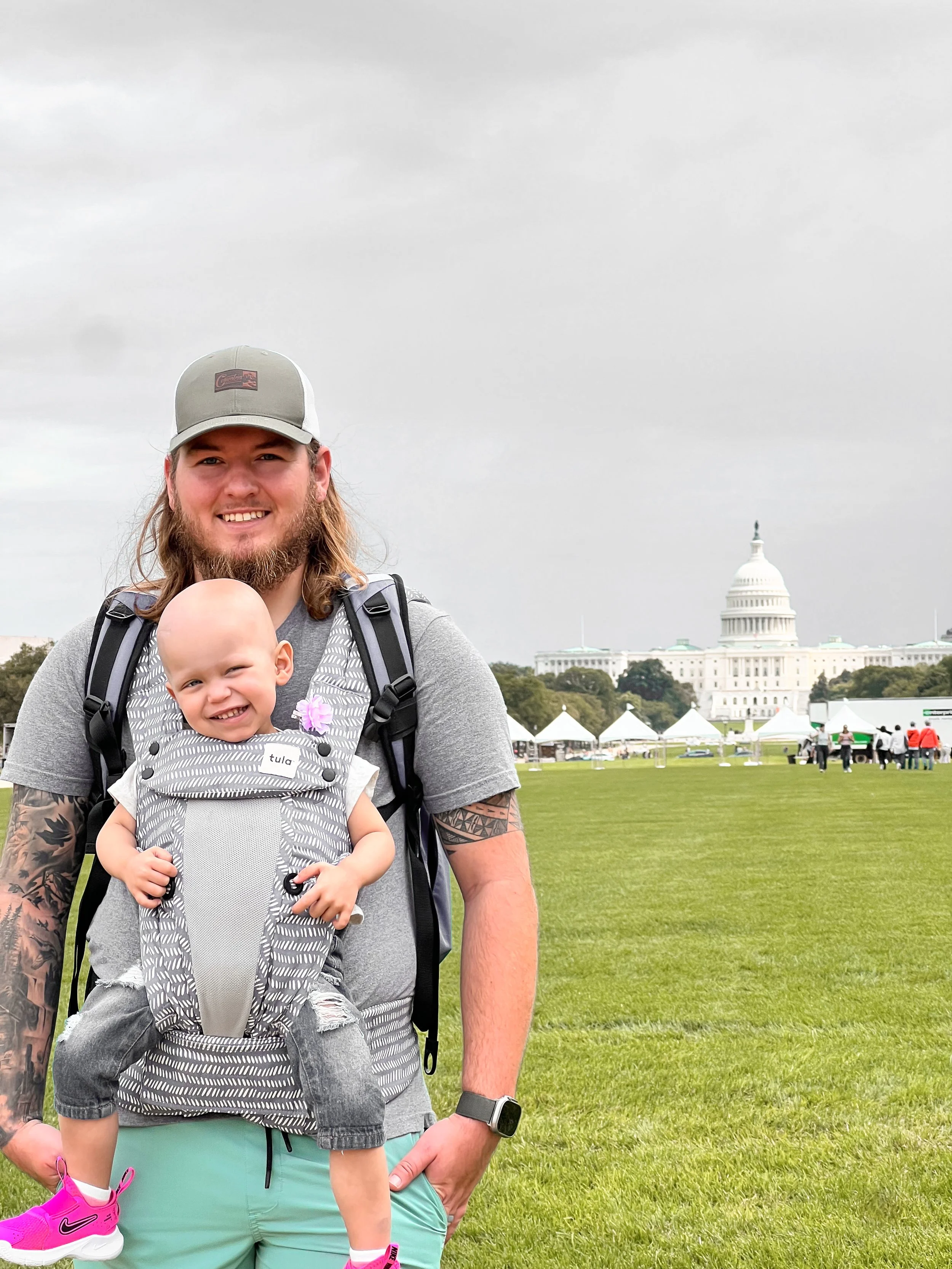 A man with long hair, a beard, tattoos, wearing a gray T-shirt, a gray cap, and a smartwatch, smiling, carrying a young girl in a gray and white baby carrier. The girl has a flower in her hair and is wearing a gray outfit with pink shoes. They are outdoors on a grassy area with tents and the U.S. Capitol building in the background under cloudy skies.