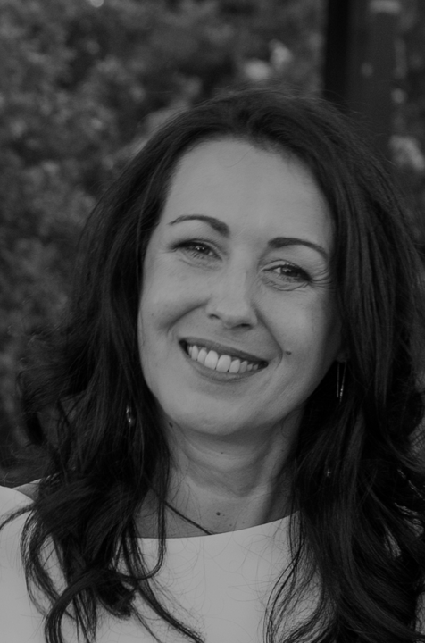 Portrait en noir et blanc d'une femme souriante avec des cheveux longs et ondulés, portant un haut clair, extérieurement dans un environnement naturel.