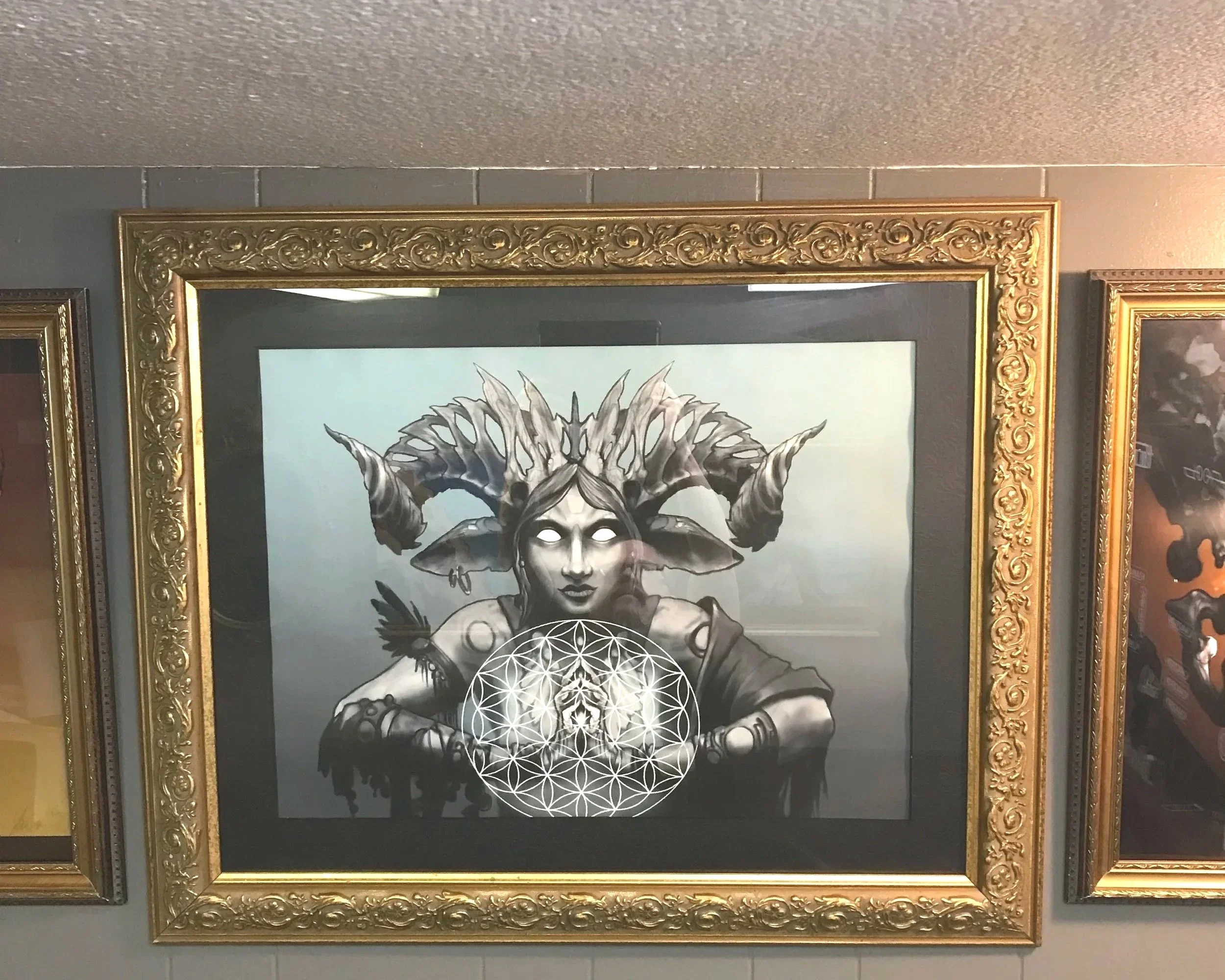 A framed black and white artwork of a mythical creature with a woman's face, crown, and antlers, with geometric patterns beneath her hands.