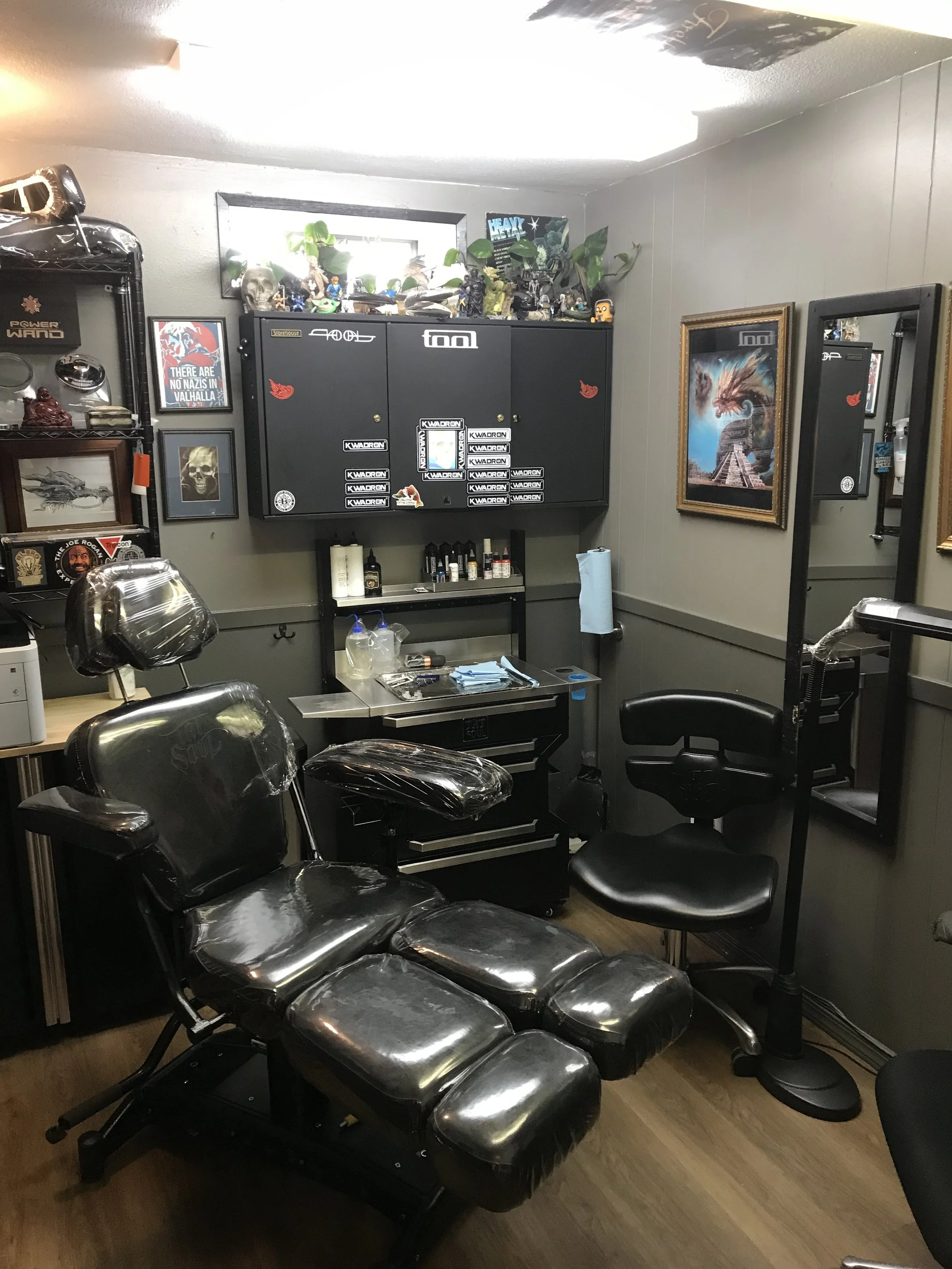 A room with multiple black massage chairs covered in plastic, a black cabinet with stickers on it, a small table with bottles and papers, framed artwork on the walls, a mirror, and various decorations including plants and posters.