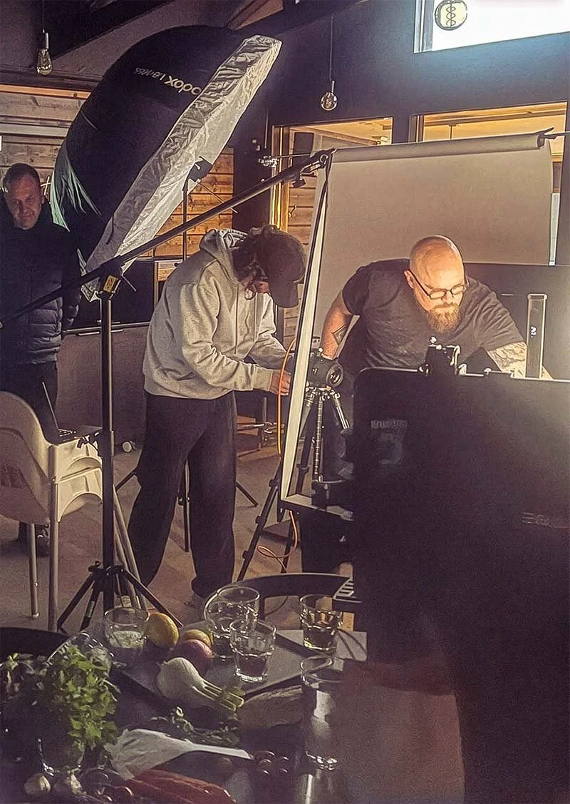 Valokuvausstudiossa kolme ihmistä työskentelee valokuvauksessa. Taustalla takki ja valo, pöydällä juomia ja hedelmiä.
