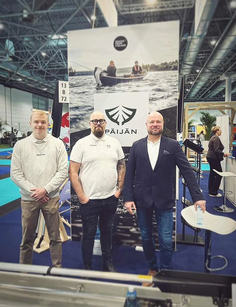 Kolme miestä seisoo messuosaston edessä, taustalla juliste ja osasto, jossa on veneitä ja muita kalusteita.