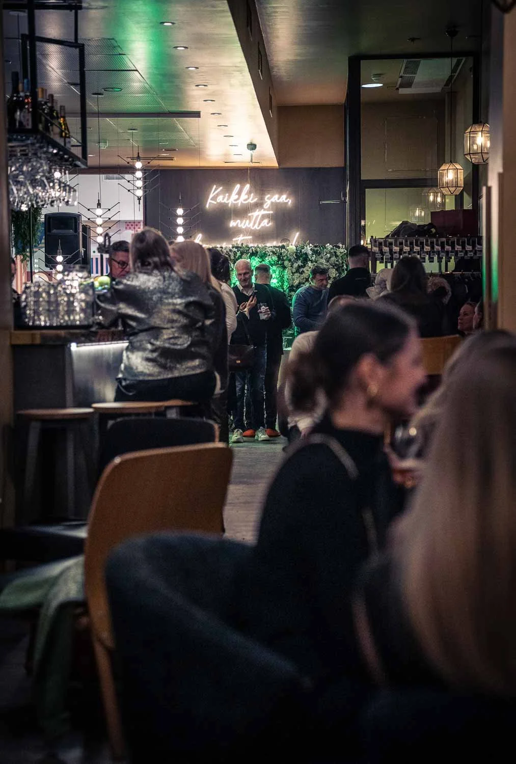 Rakennus sisällä baarista tai ravintolasta, monia ihmisiä seurustelemassa, taustalla valotaulu jossa lukee 'Kaikki saa, mutta komiat ensin'.