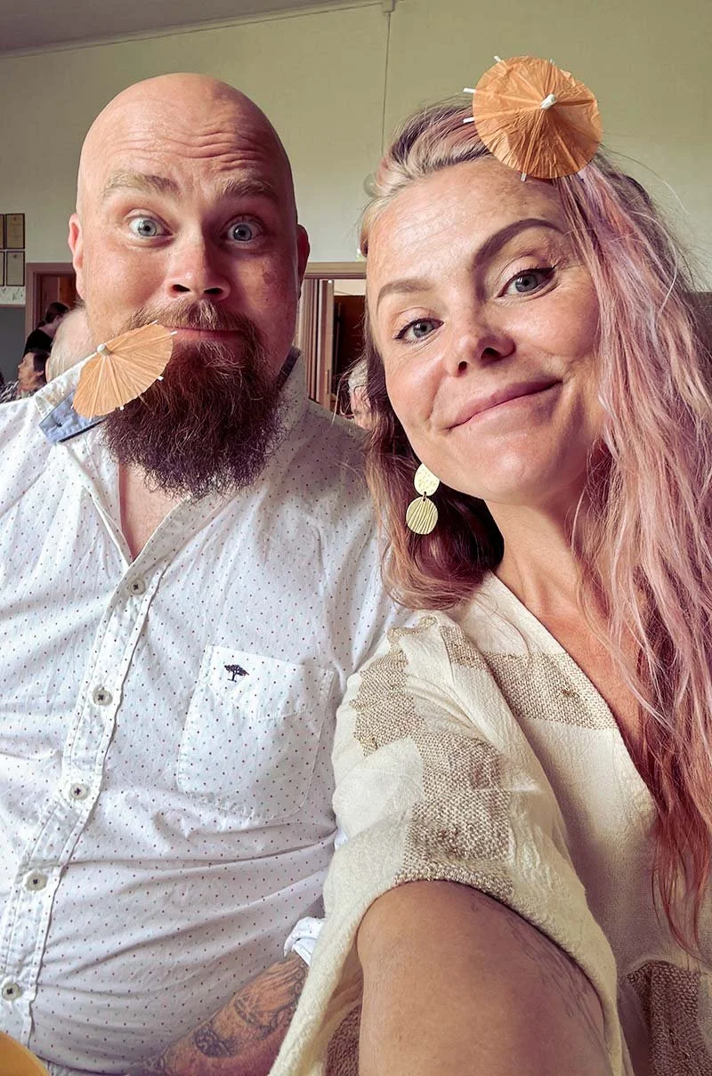 Kaksi ihmistä poseeraa selfie-valokuvassa, joilla on pieni paperinen cocktail- tai paperipilli tuotteen suussa, ja toinen heistä käyttää jättimäisiä koruja sekä päässään pieni paperinen hattunen.