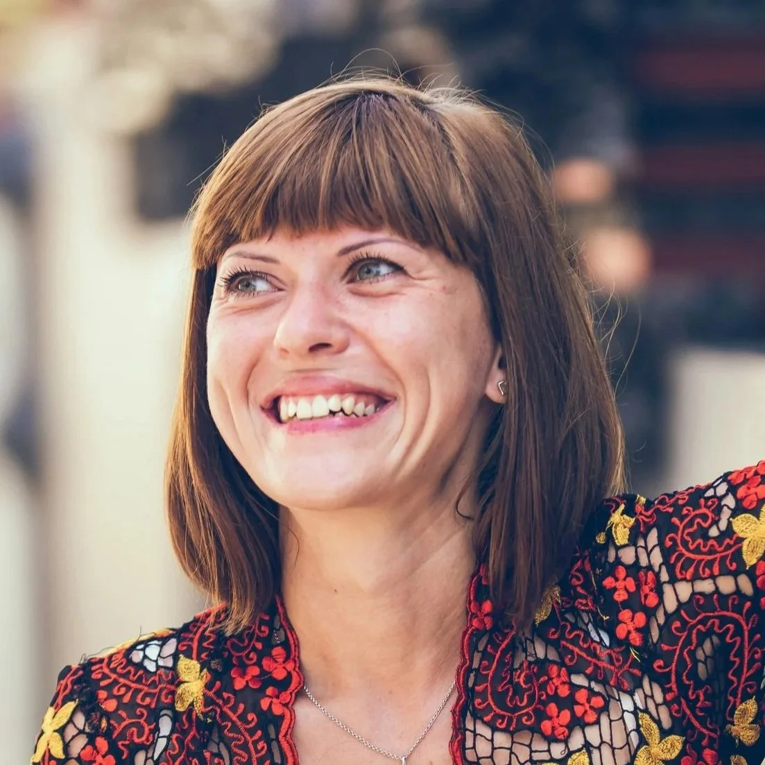 A woman with shoulder-length brown hair and bangs, smiling and looking slightly to her right, wearing a black top with colorful embroidered floral patterns.