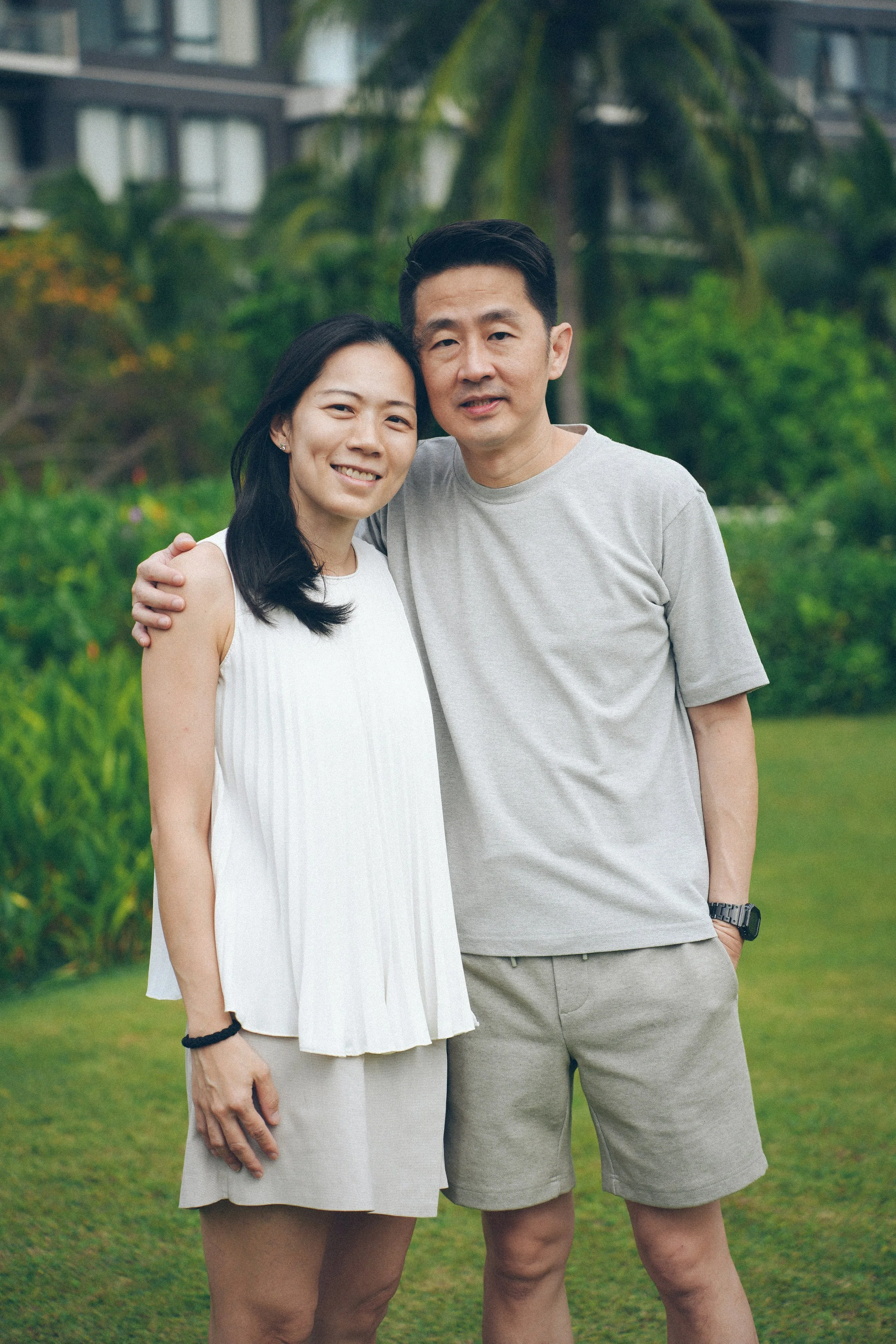 A smiling woman and man standing together outdoors, with the woman resting her head on the man's shoulder. They are in a green park with trees, bushes, and a building in the background.