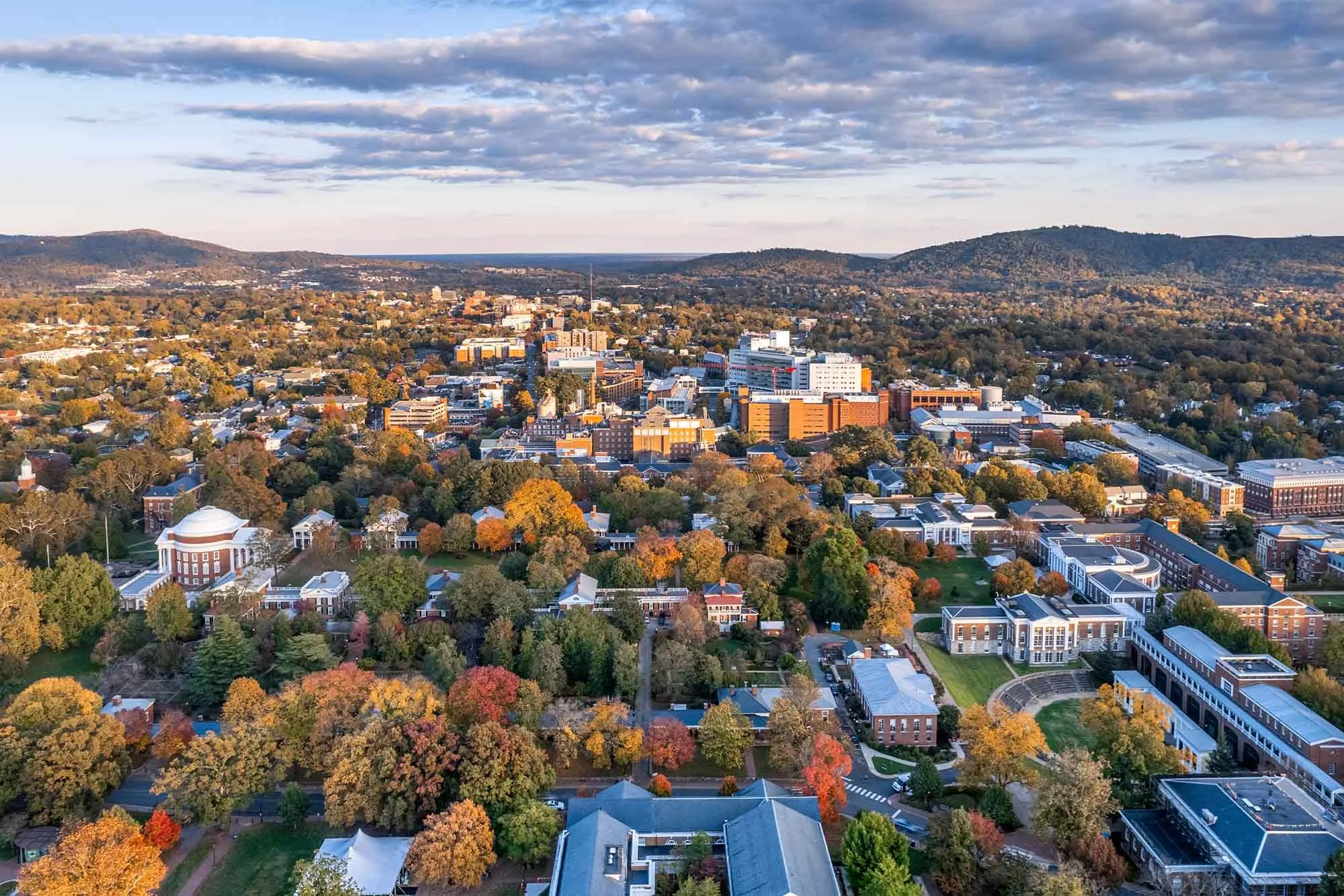 Aerial view of the University of Virignia campus in Charlottesville in autumn, showing trees with colorful fall foliage, and buildings including academic halls and administrative offices surrounded by hills and mountains.