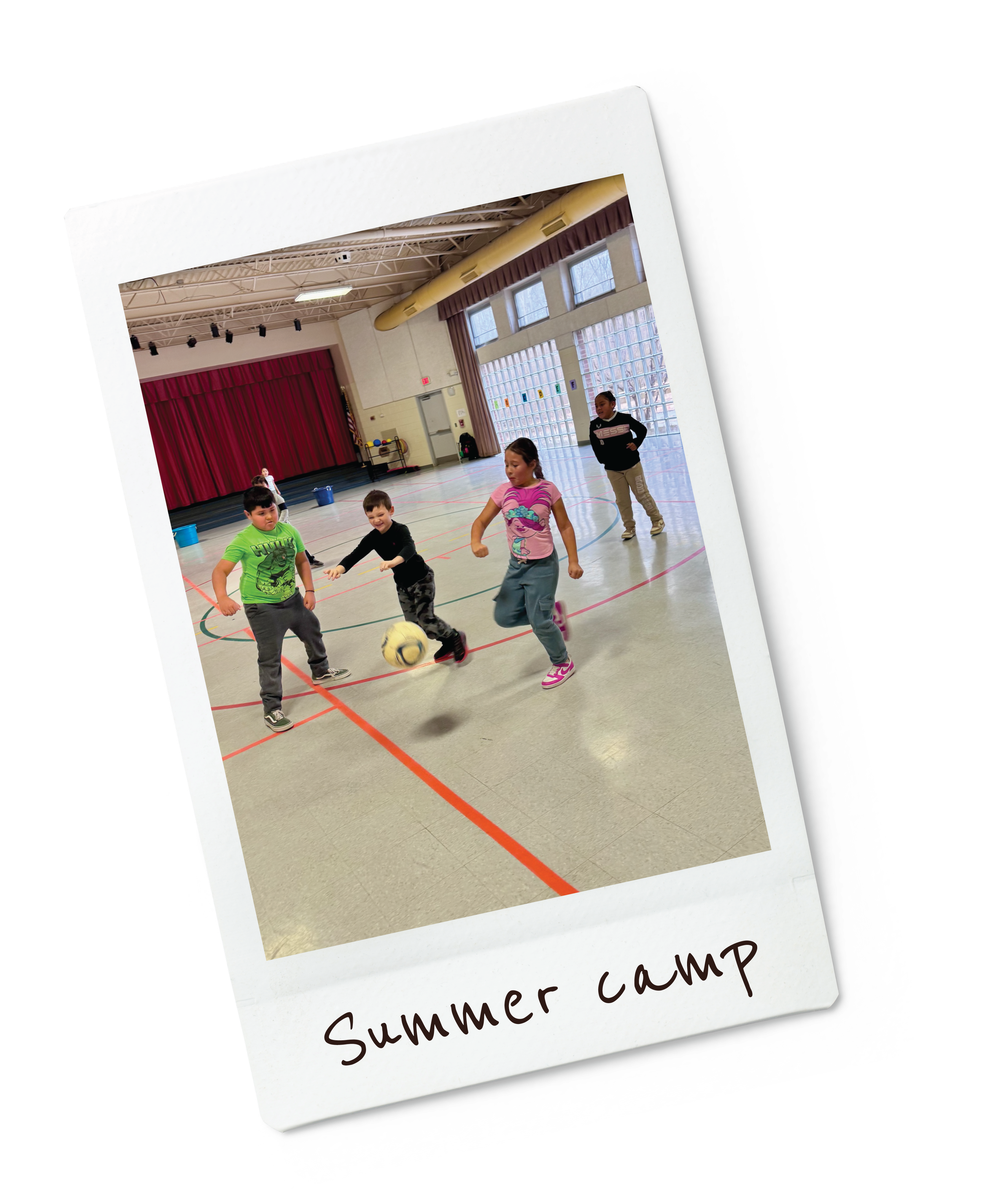 Kids playing soccer indoors at summer camp, gymnasium with red curtains, large windows, and high ceiling.