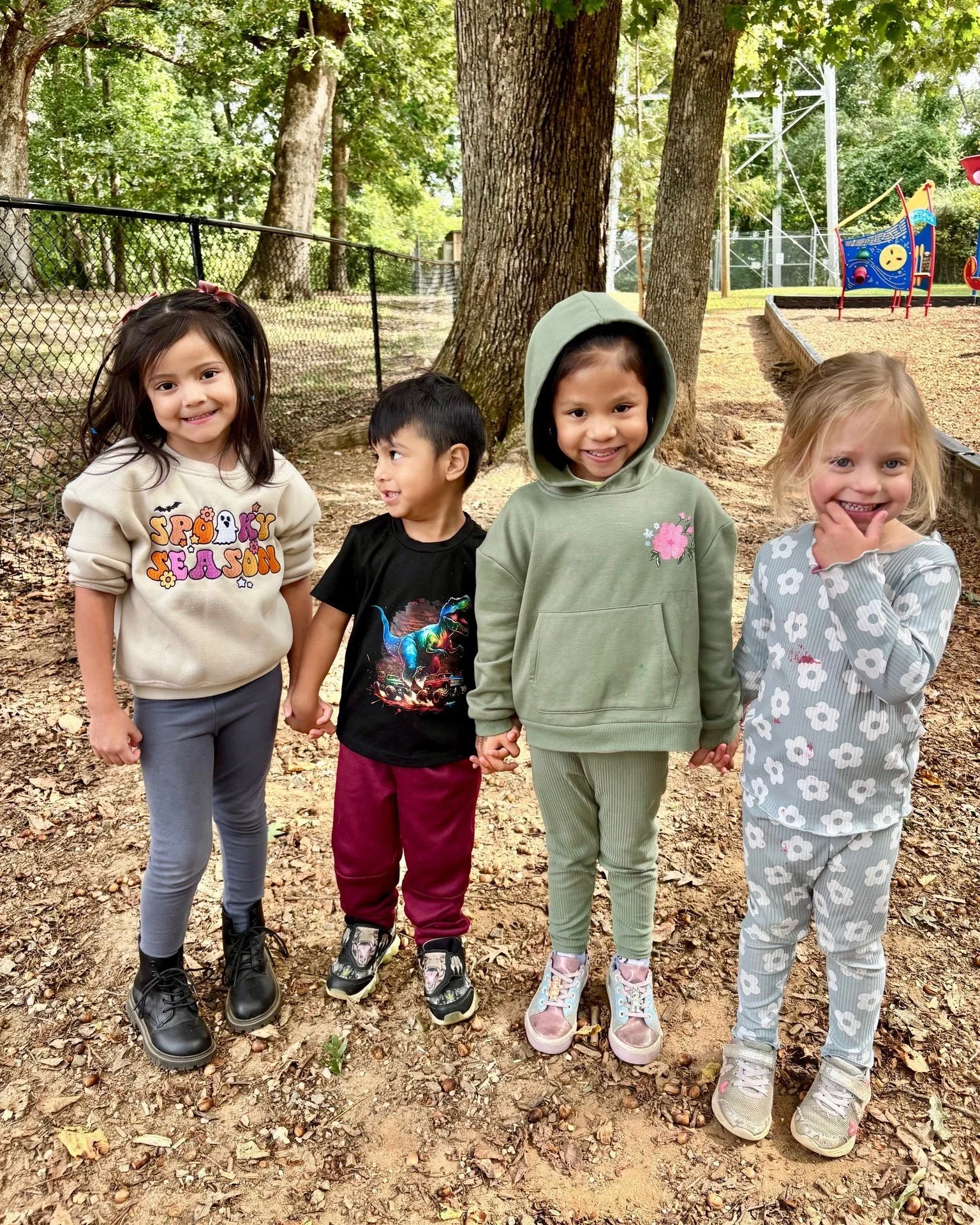 Four happy children holding hands outdoors in a wooded park with trees and playground equipment in the background.