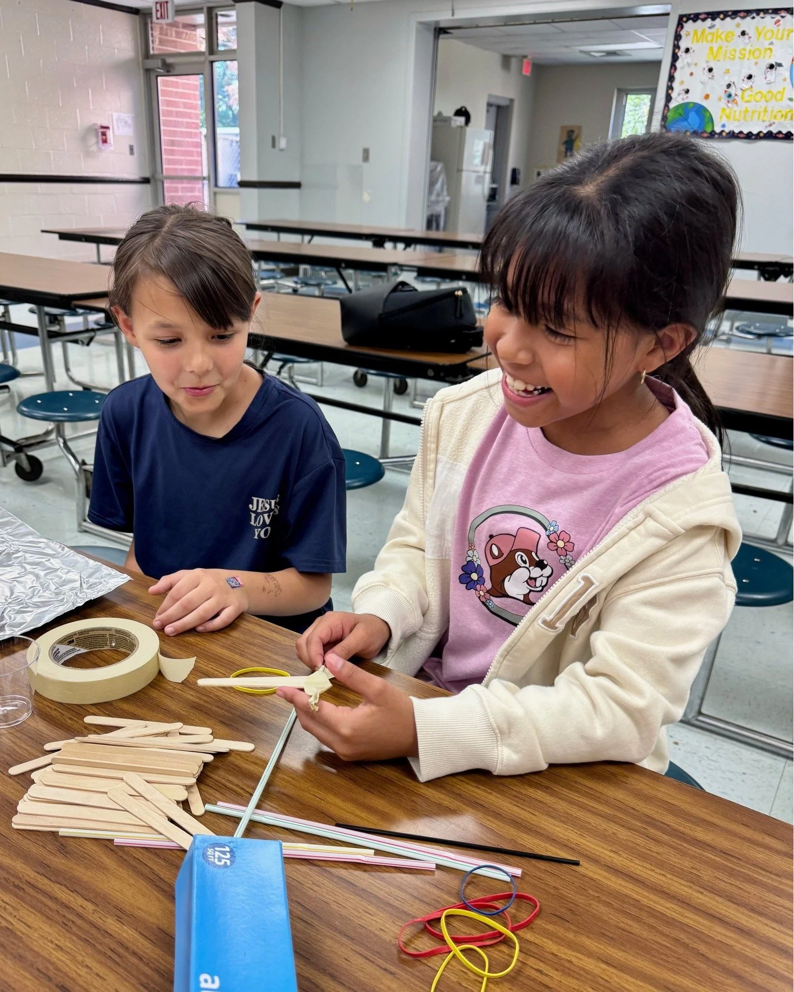 Two children, a boy and a girl, sitting at a table in a classroom. The girl is holding a popsicle stick with a rubber band on it, smiling. They are surrounded by other popsicle sticks, tape, and rubber bands.
