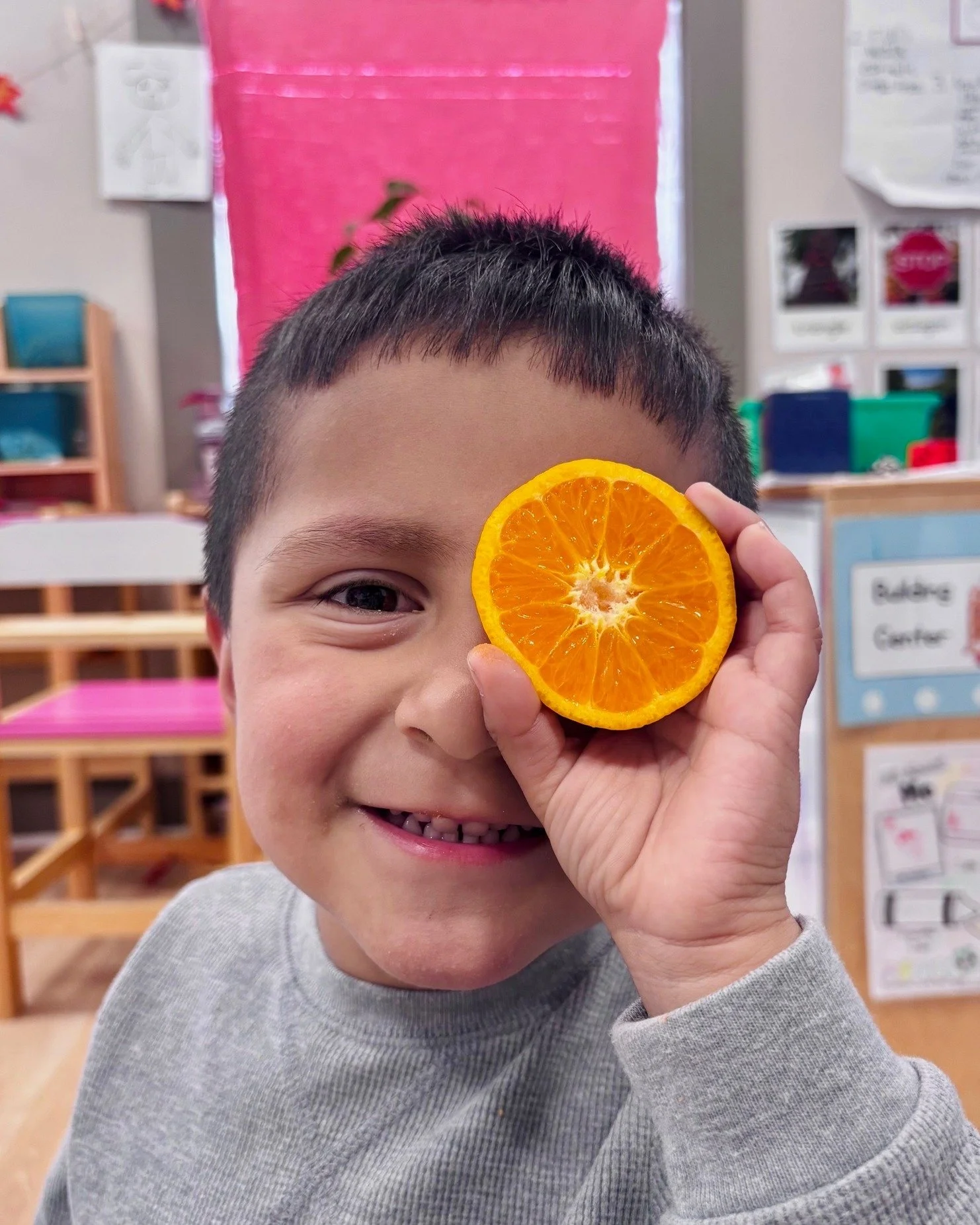 A young boy smiling and holding a slice of orange over his eye in a classroom setting.