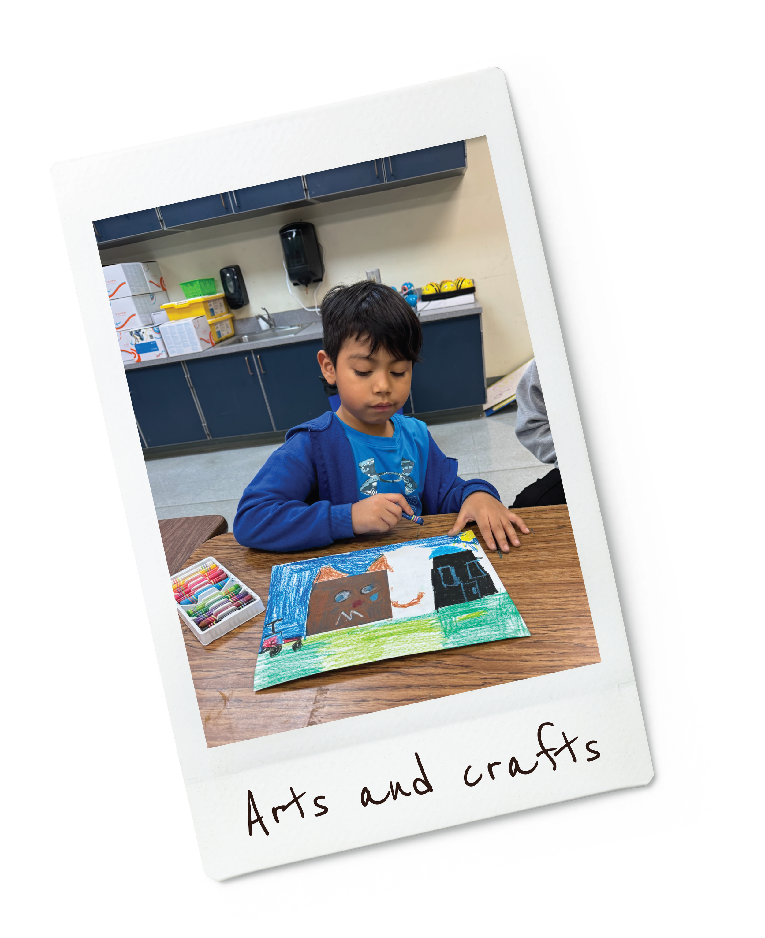 A young boy in a blue shirt sits at a table drawing a colorful picture with crayons. The photo has a label "Arts and crafts." The background shows a school or community center kitchen with cabinets, a sink, and various supplies.