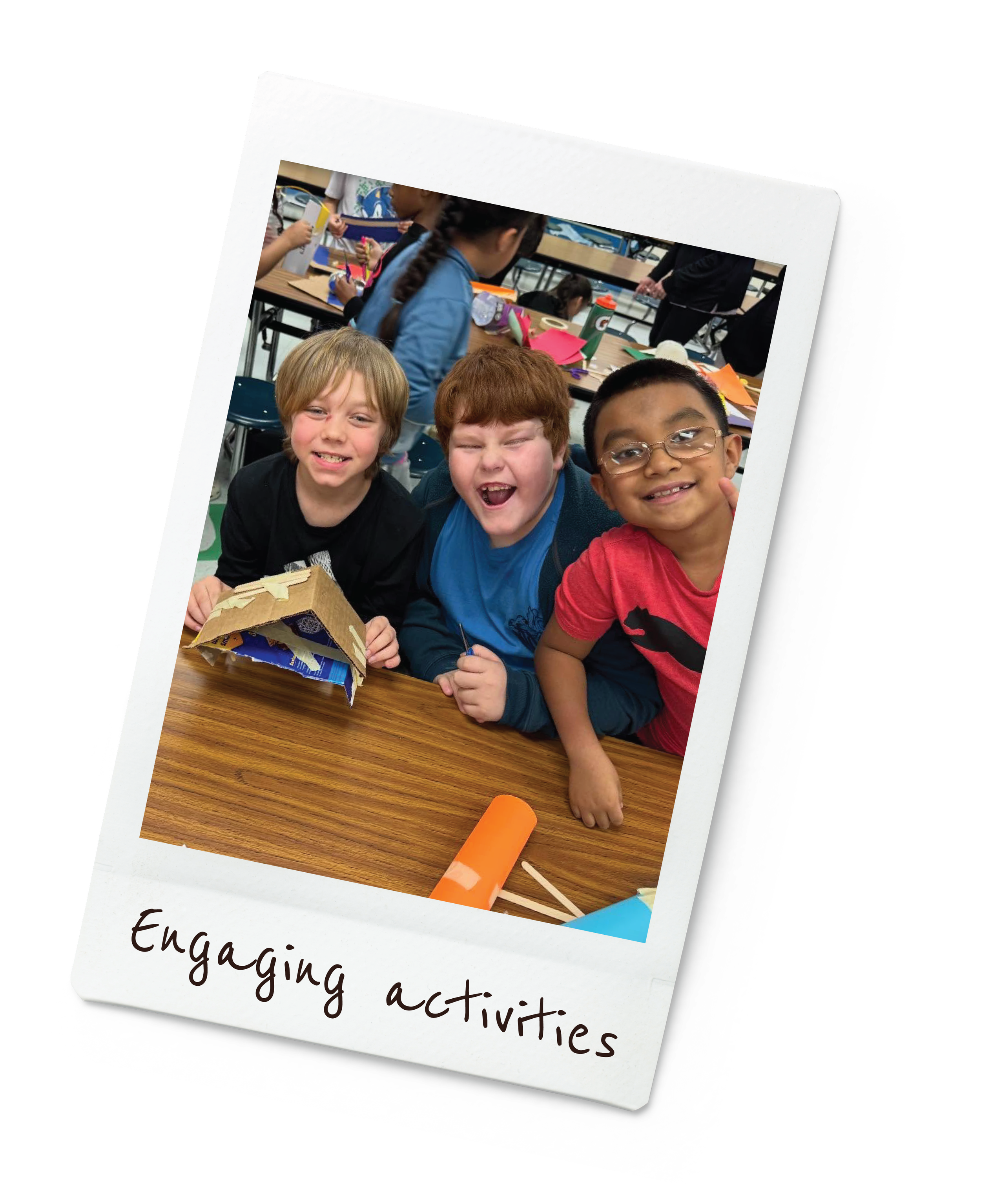 Three kids smiling and laughing at a table during a classroom activity, with craft supplies in the background.