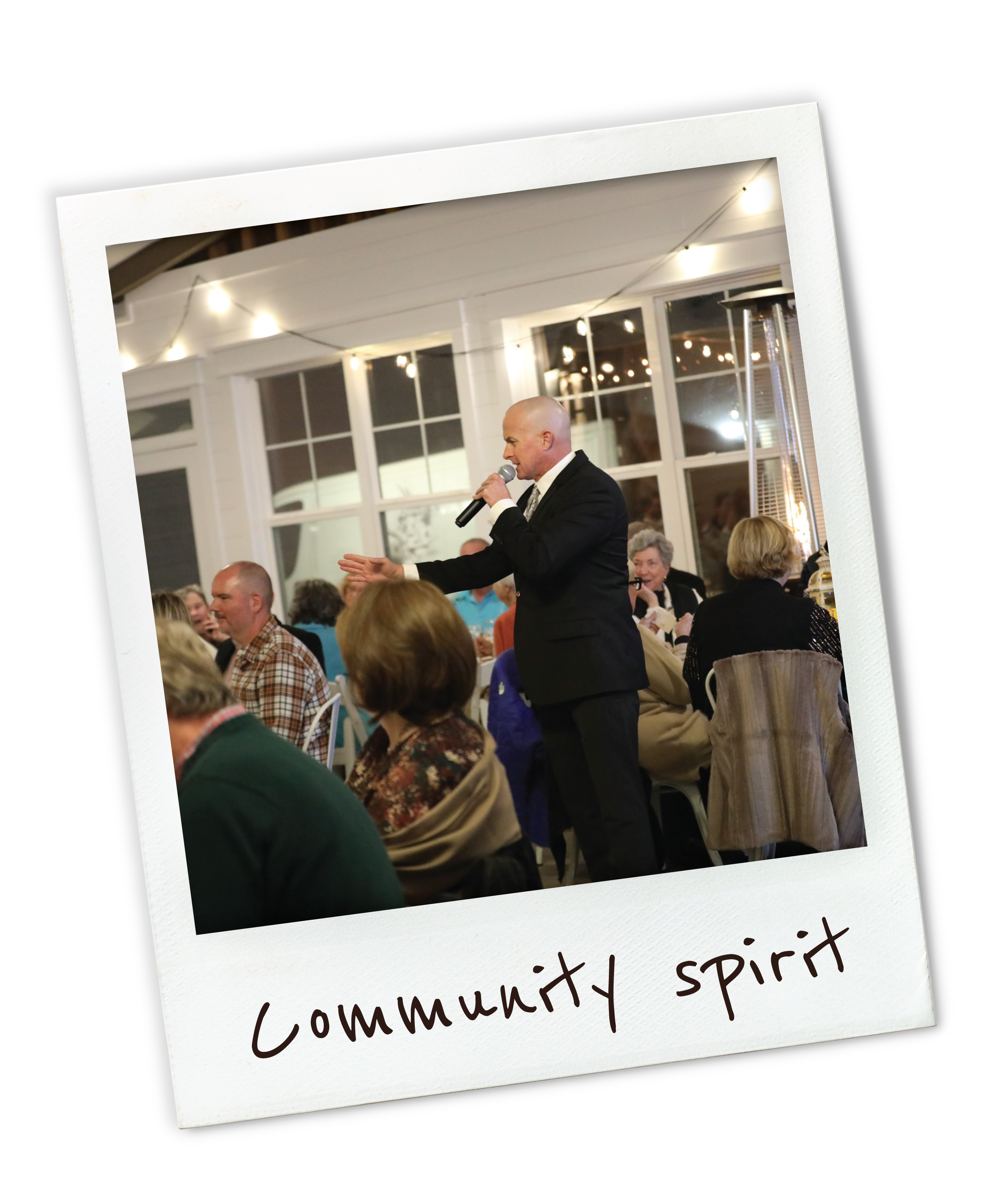 A man in a black suit speaking into a microphone at a community event, with an audience seated at tables, in a well-lit, indoor space with string lights and large windows.
