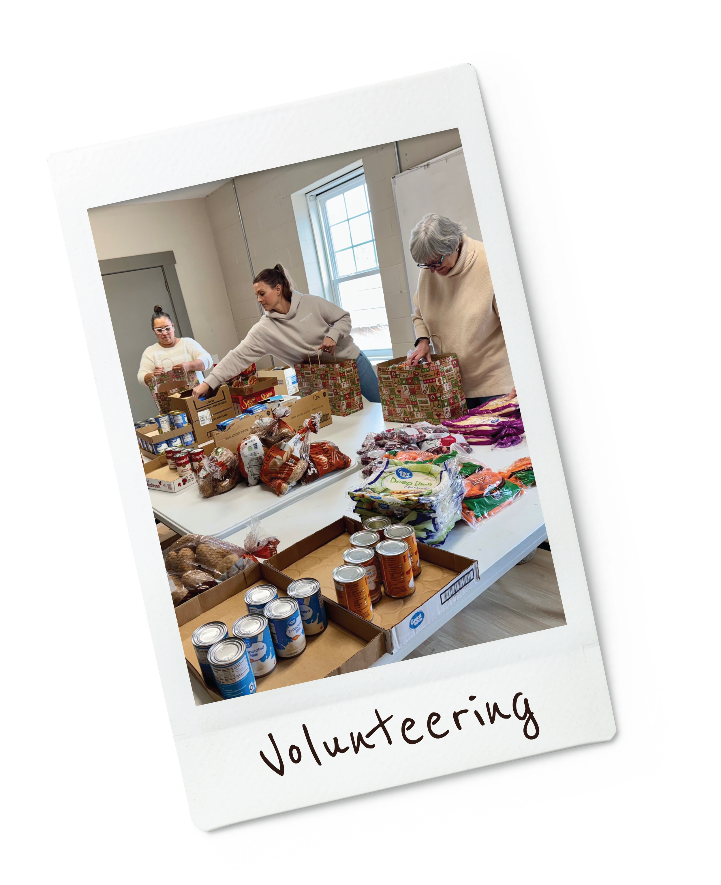 Three women volunteer packing food items into boxes at a table during a charity event, with canned goods, produce, and packaged food on the table, in a room with a window and white walls.