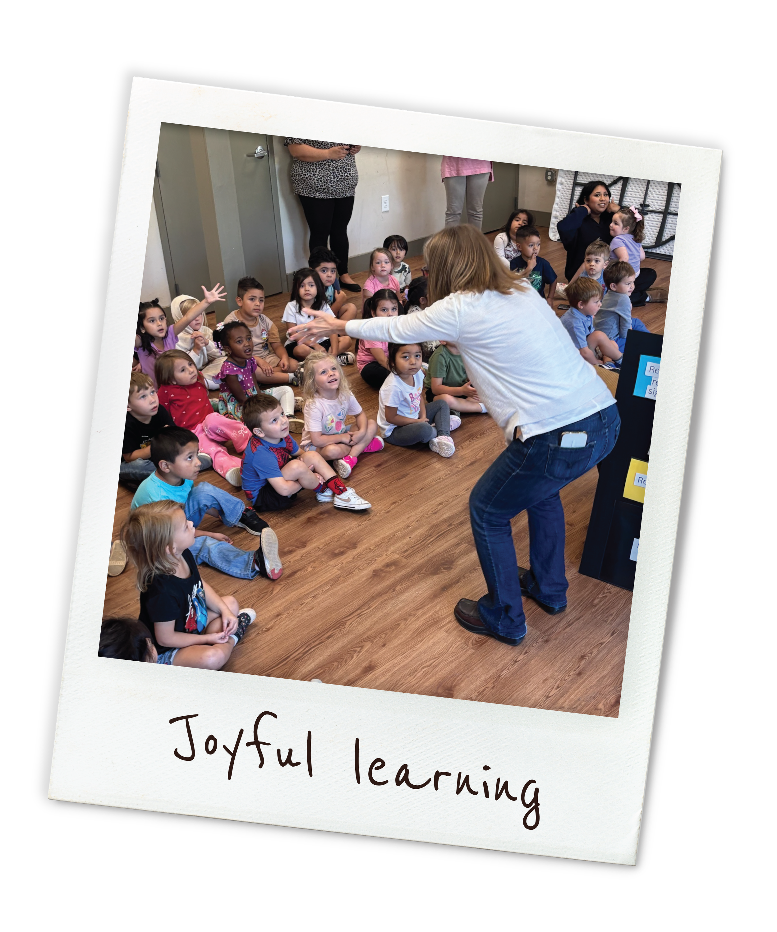 A woman leading a group of young children seated on the floor during a classroom activity, with some children raising their hands, and two other adults standing in the background. The photo is framed as a Polaroid with the caption "Joyful learning."