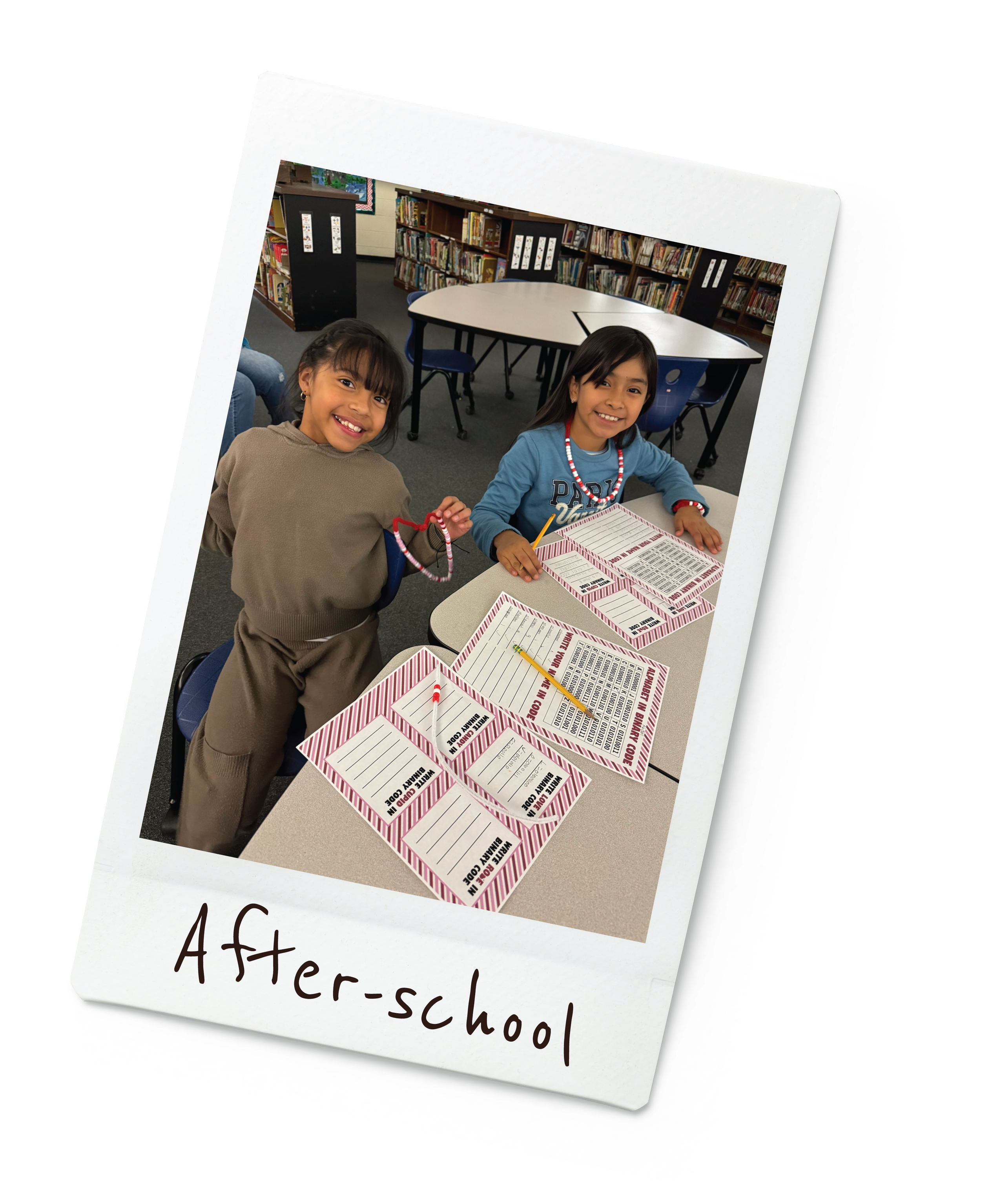 Two young girls smiling and sitting at a table in a library, completing reading or activity worksheets with pencils and reward charts. One girl is wearing a tan outfit, and the other is wearing a blue shirt with beaded necklaces. The photo has the caption 'After-school' at the bottom.