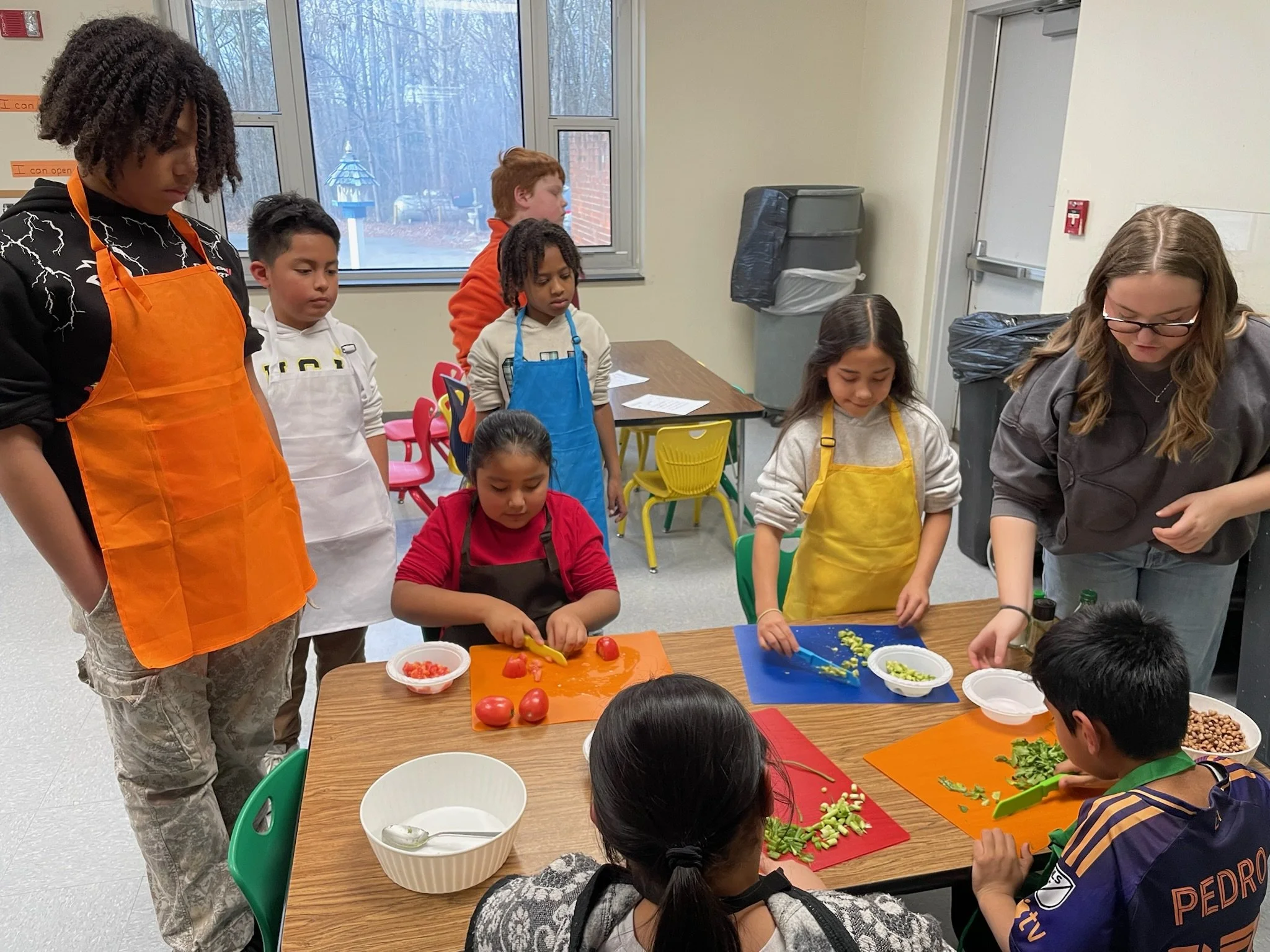 Group of children and instructors preparing food with vegetables on colorful cutting boards in a classroom.