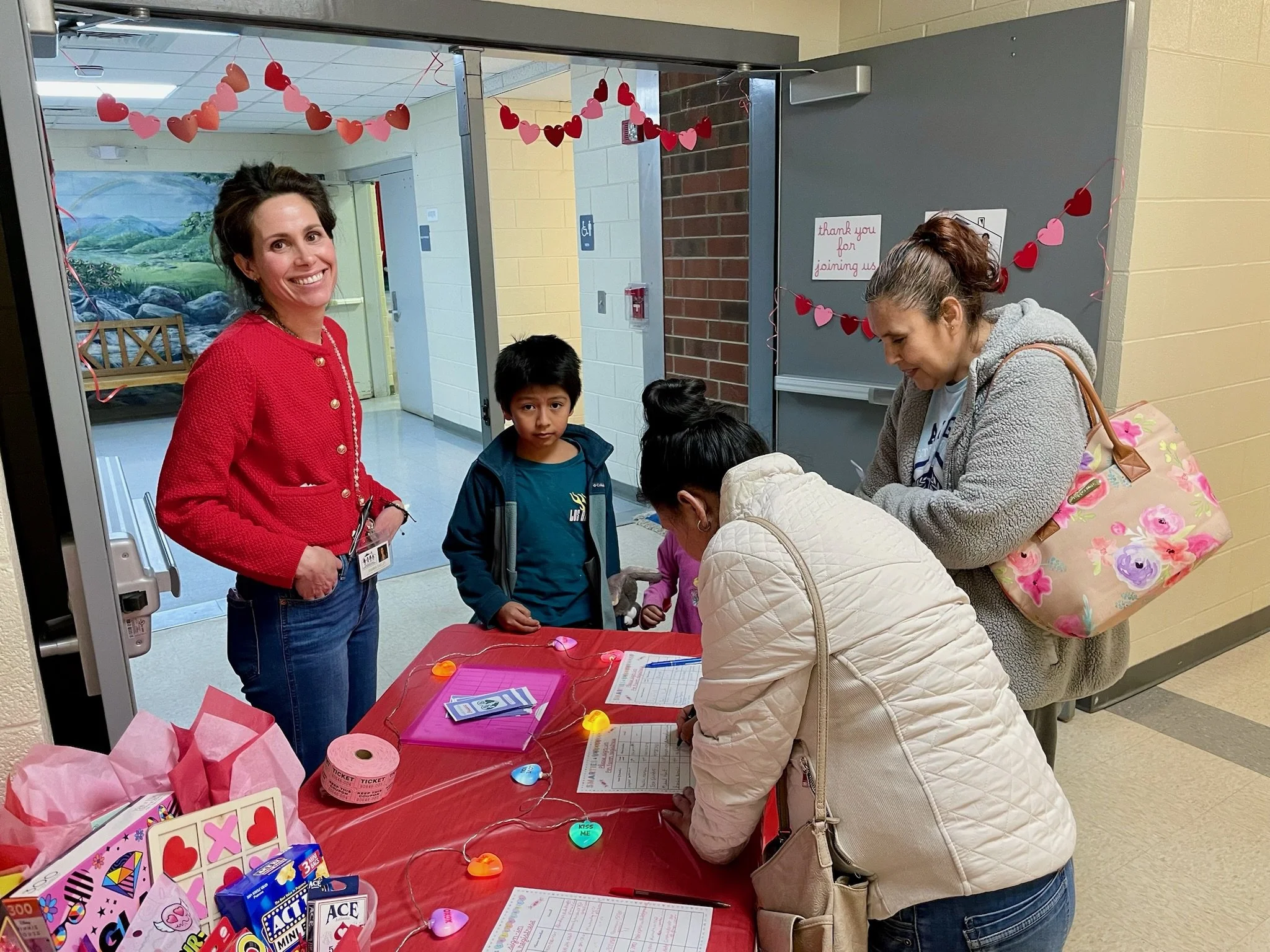 A group of people at a decorated table in a school hallway, with Valentine’s Day decorations and a woman in a red jacket smiling.