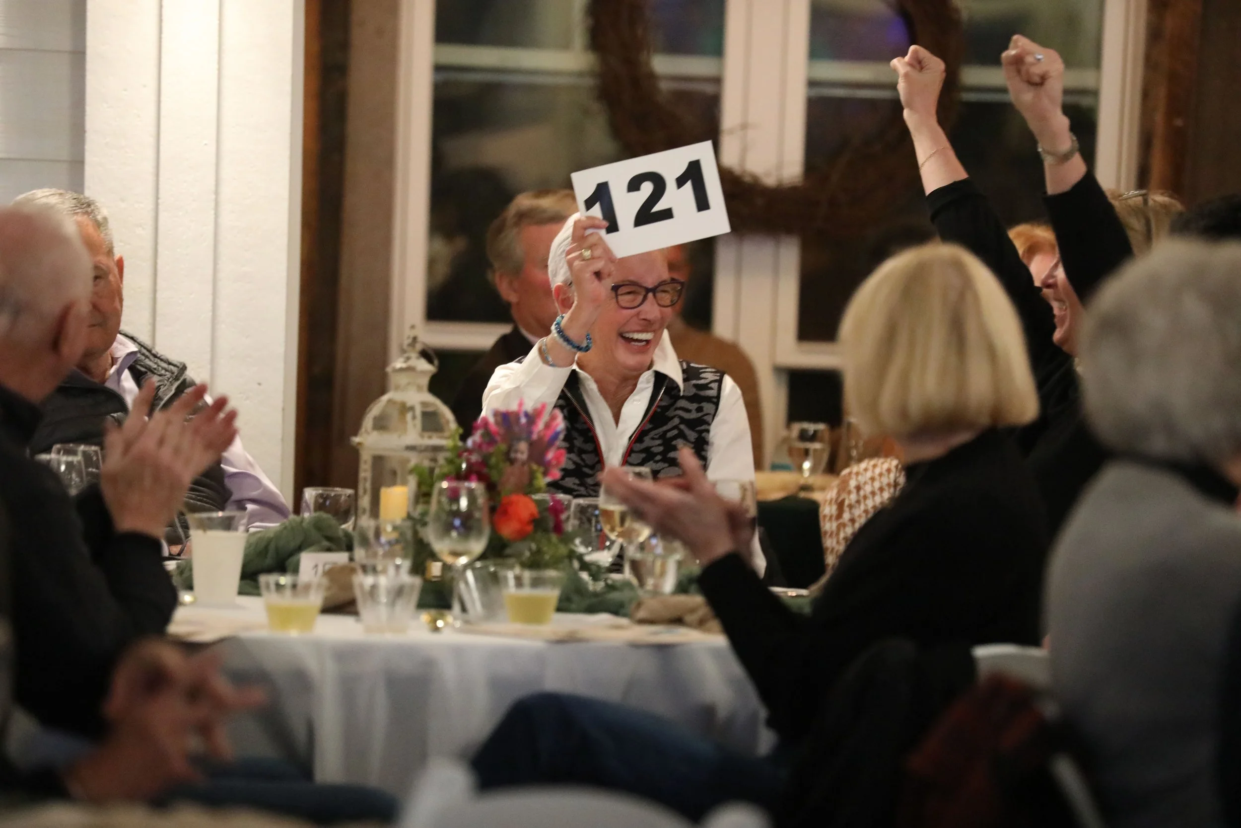 A woman at a dinner celebration holding a sign with the number 121, smiling and celebrating with others in a festive atmosphere.
