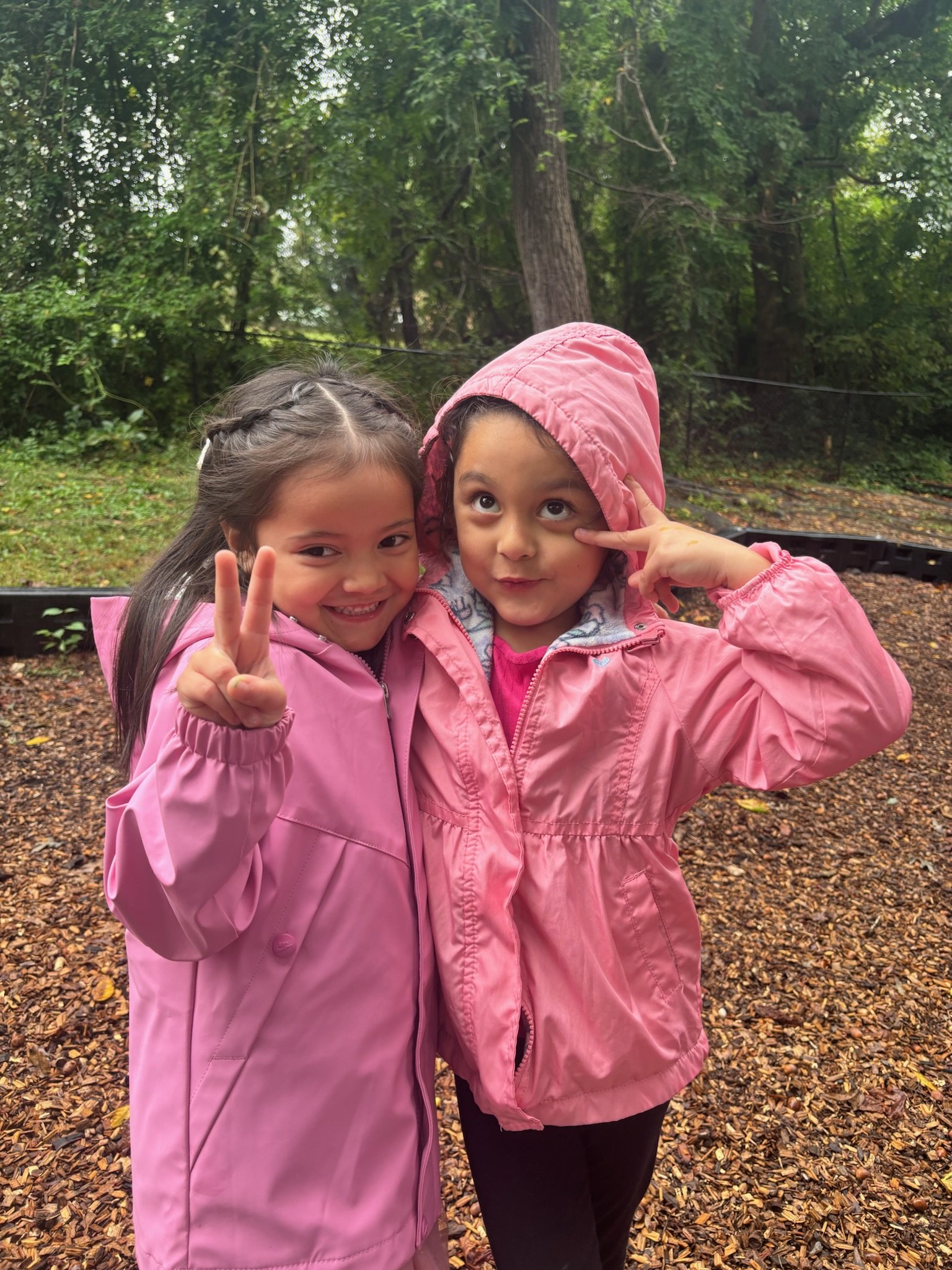 Two young girls in pink jackets and rain hats standing outdoors in a wooded area, smiling, with one girl showing a peace sign and the other making a playful gesture near her eye.