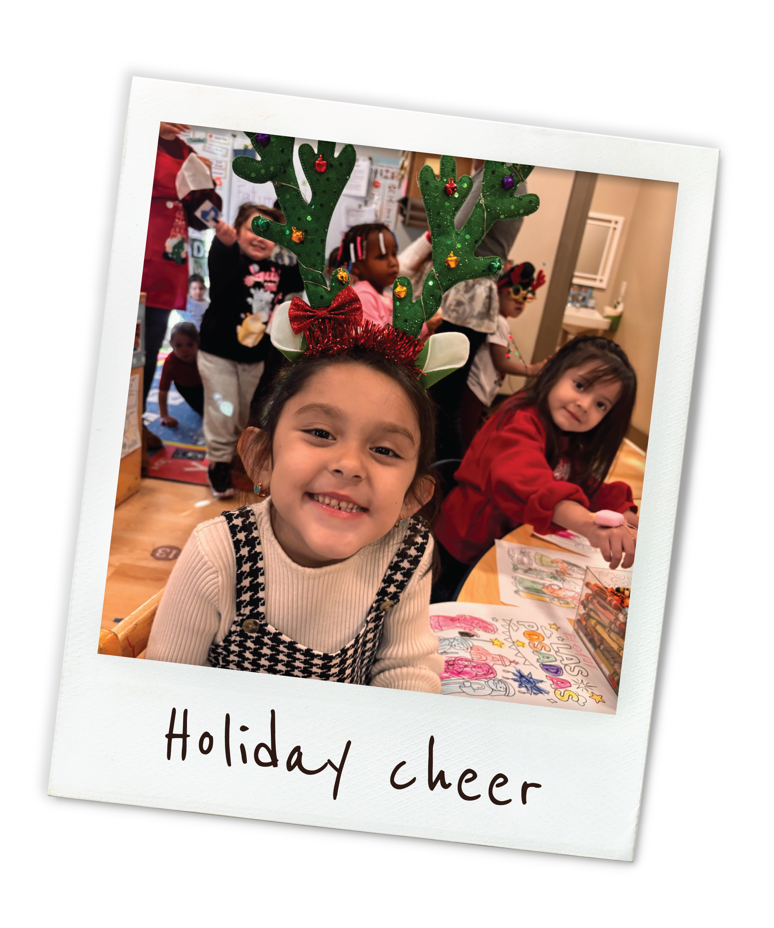 Child wearing reindeer antlers headband smiling at the camera during a holiday celebration with other children in the background.