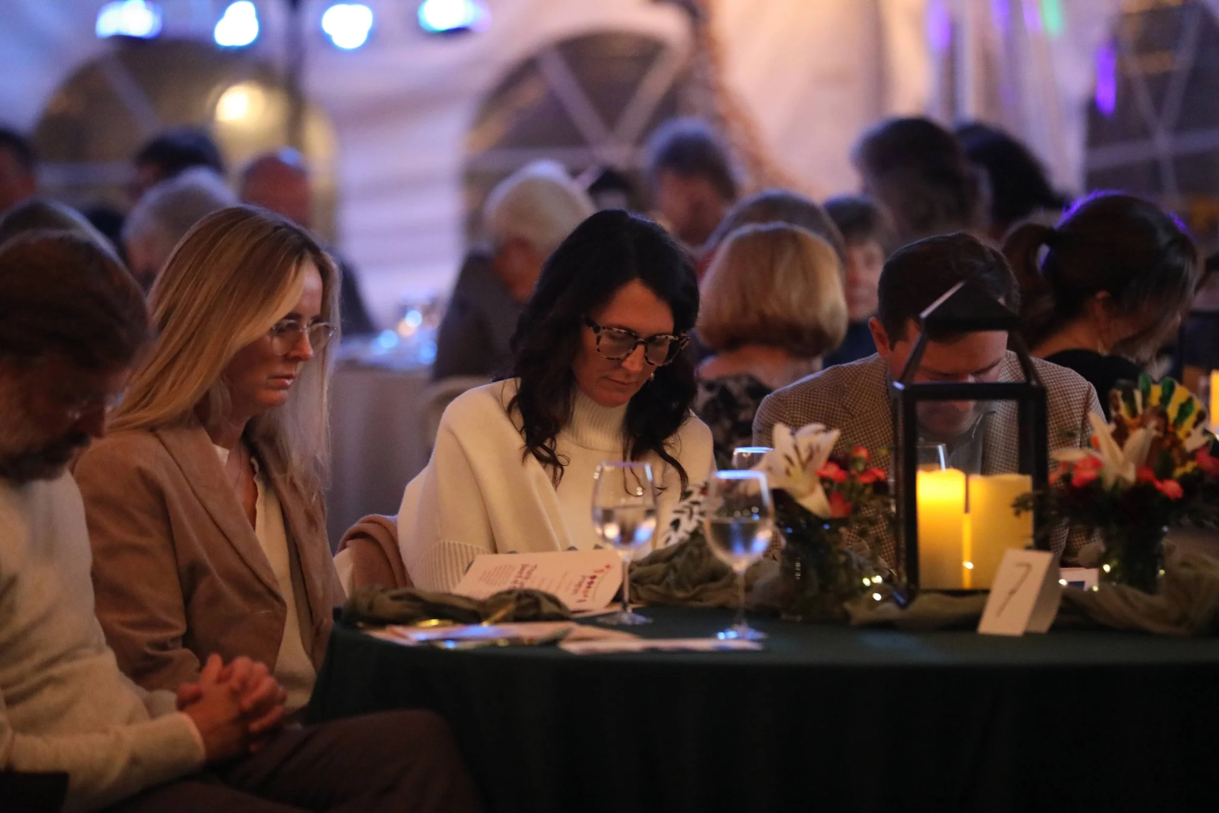People sitting at a dimly lit banquet table during an event or dinner, with floral centerpieces and candles, and some looking at menus.