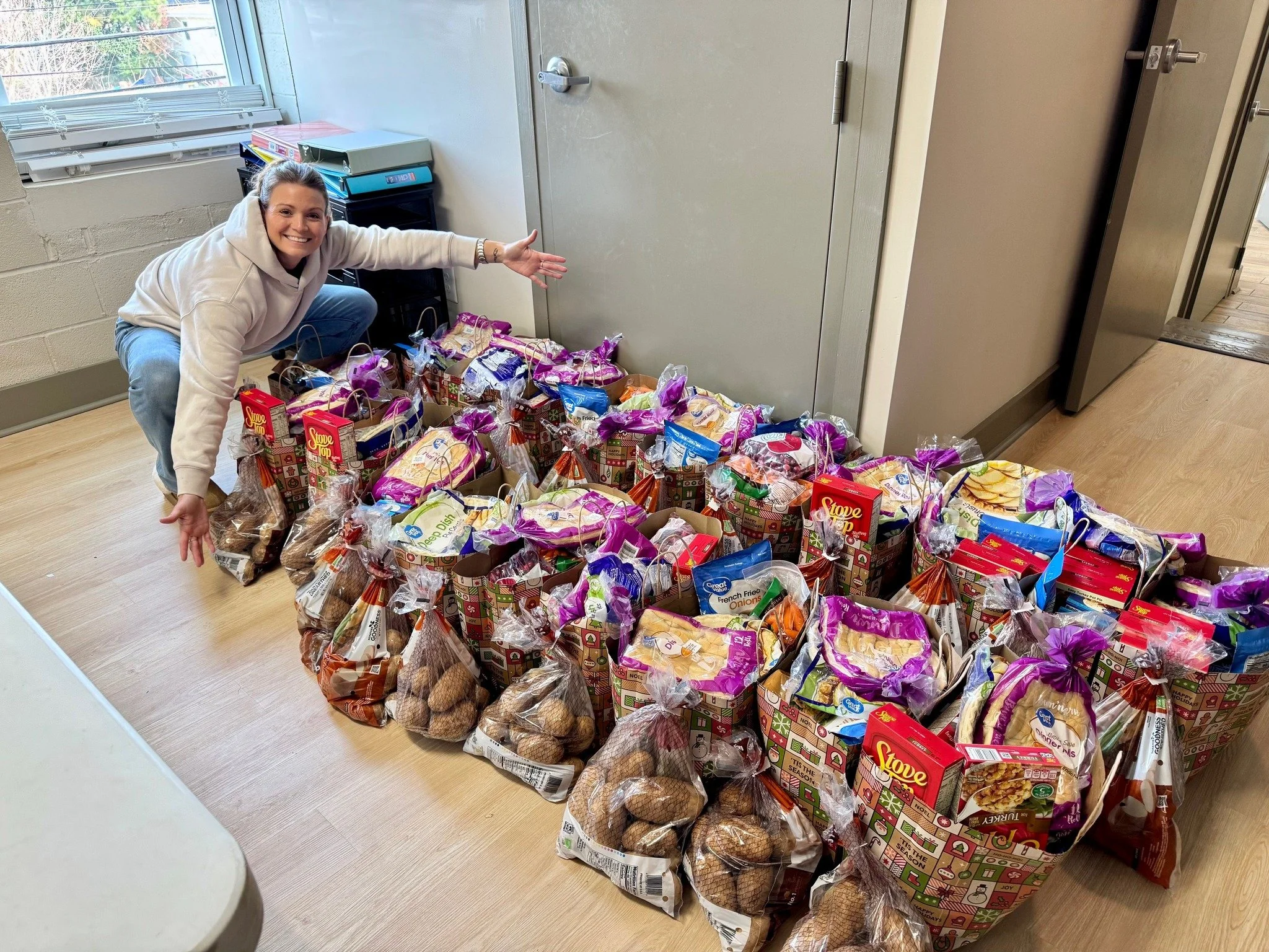 A smiling woman crouching next to a large collection of grocery bags filled with food. The bags are neatly arranged on the floor near a wall with a window. The woman is pointing towards the bags, which include potatoes, boxed food items, and other groceries.