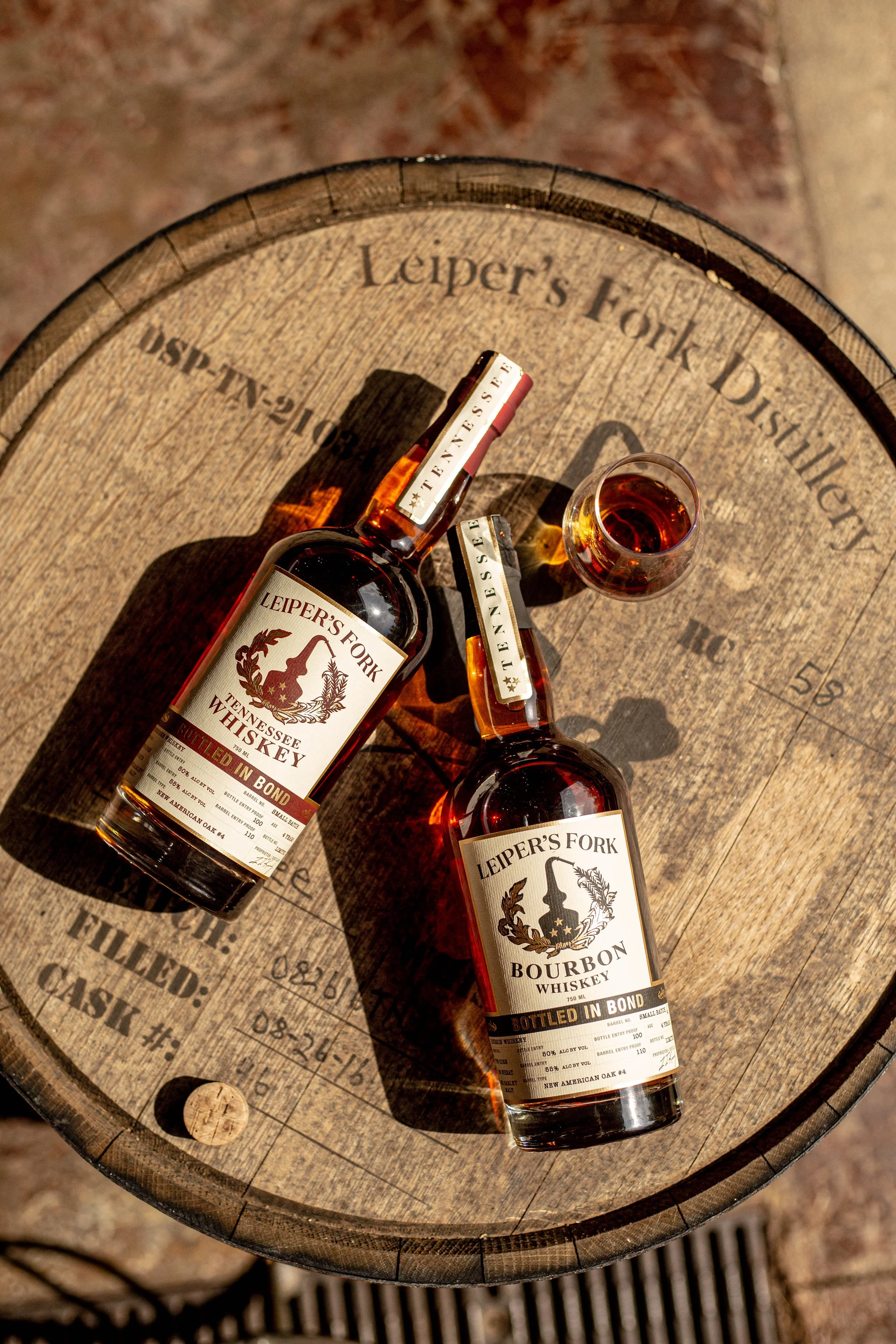 Two bottles of Leiper's Fork Tennessee whiskey, one bourbon and one whiskey, with a shot glass of whiskey on a wooden barrel top.