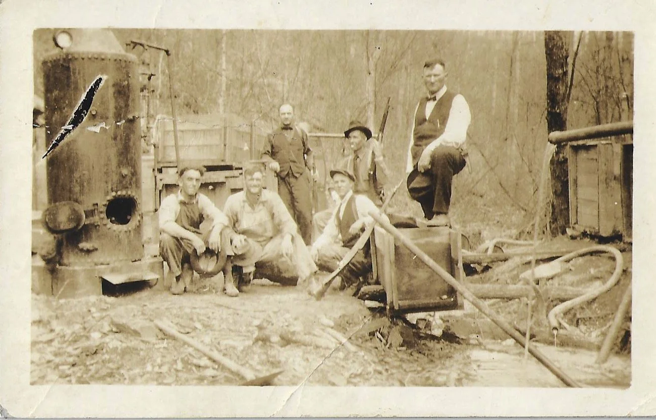A vintage black-and-white photograph of six men outdoors next to a steam engine, some wearing work clothes and hats, in a wooded area with trees and a wooden fence in the background.