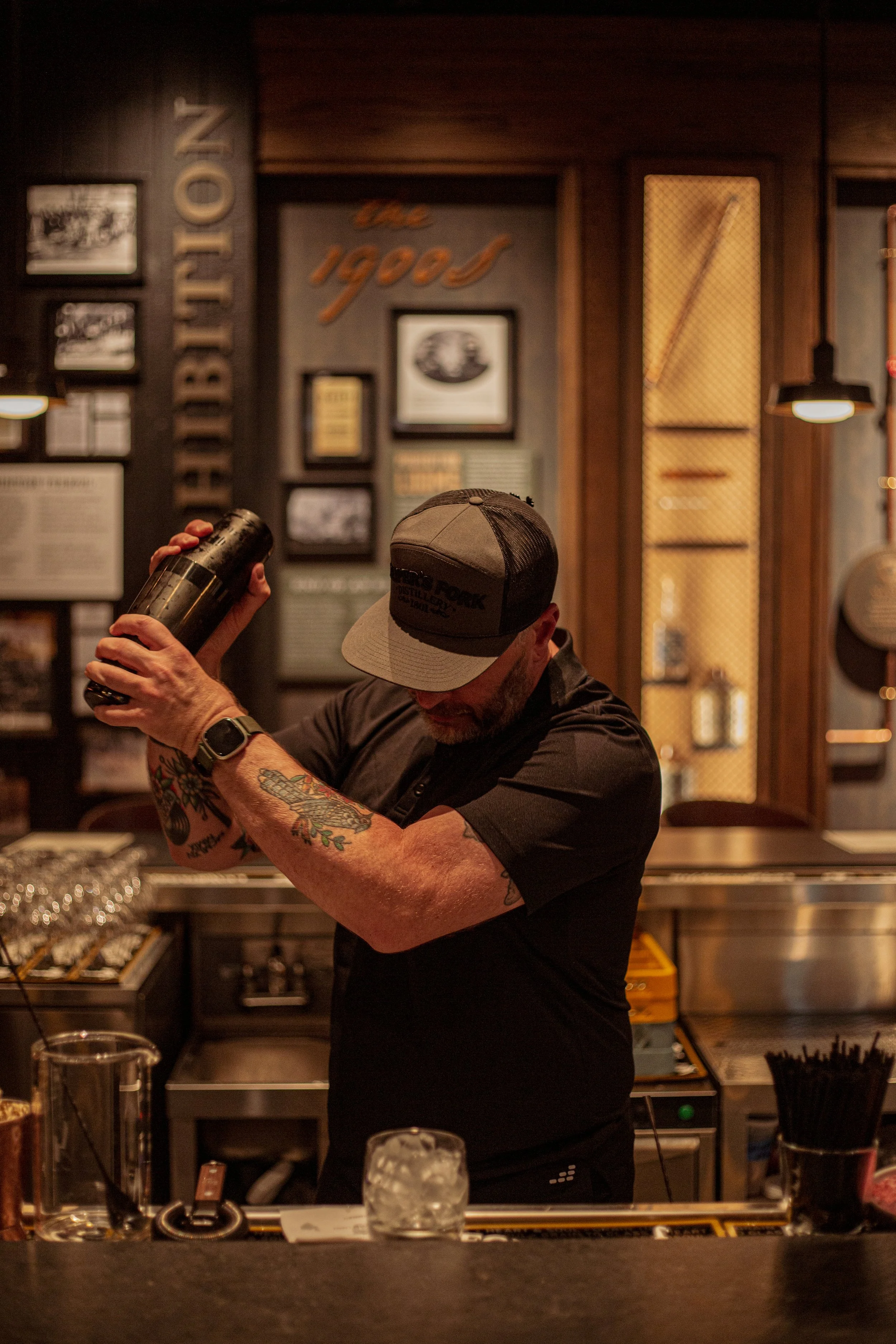 A bartender with tattoos on his arms, wearing a black shirt, a cap, and a watch, preparing a drink behind a bar in a dimly lit, rustic-style bar or restaurant.