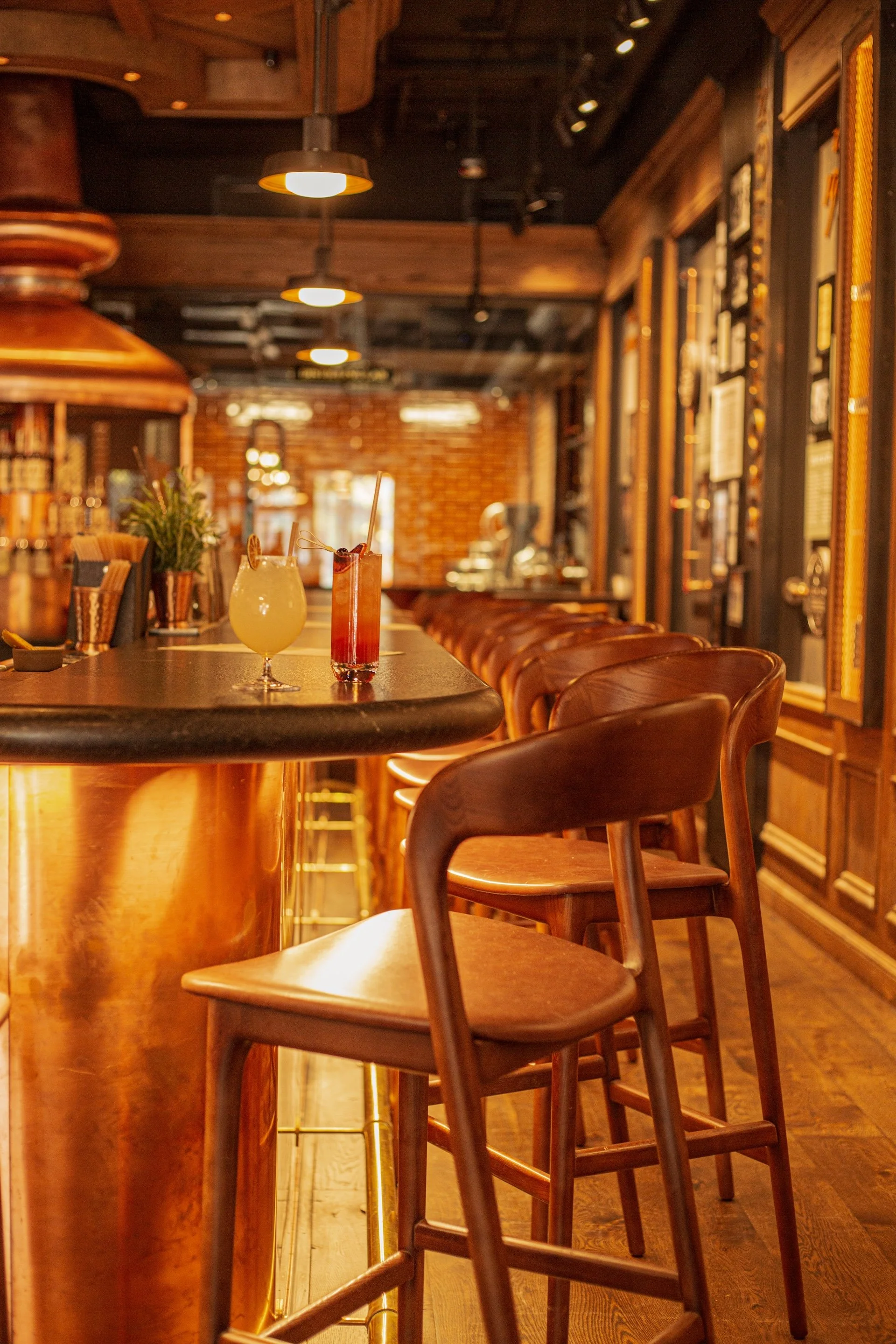 Empty bar with wooden stools and two cocktails on the counter, warm lighting, and wooden decor.