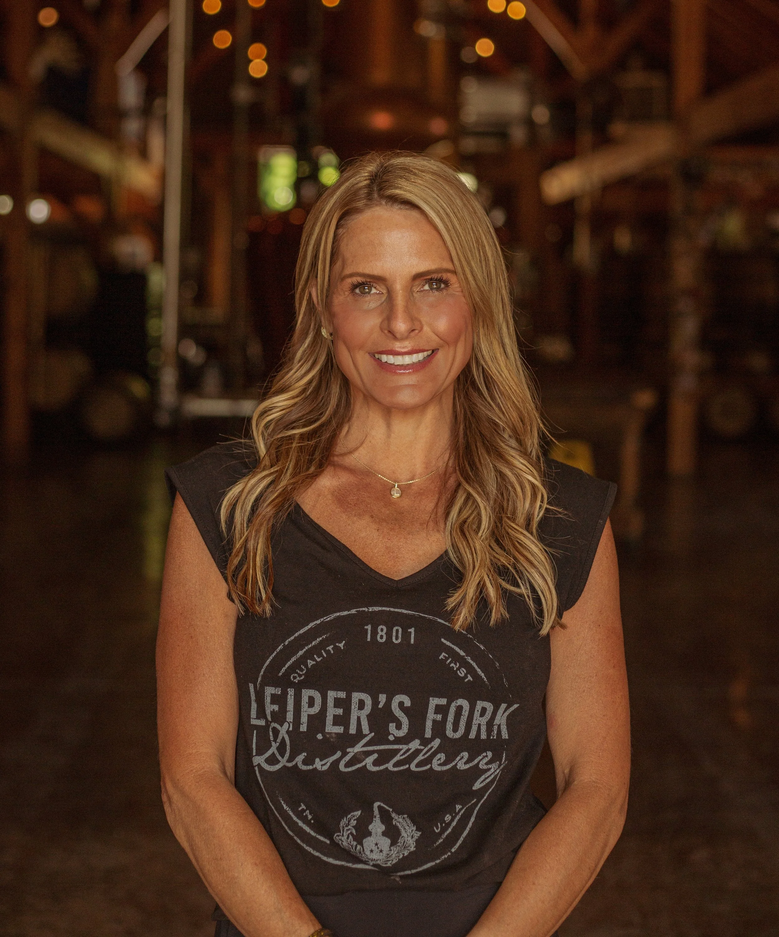 A woman with blonde hair smiling, standing inside a wooden barn, wearing a dark blue T-shirt with 'Leiper's Fork Distillery' logo and a necklace that says 'Lynee'.