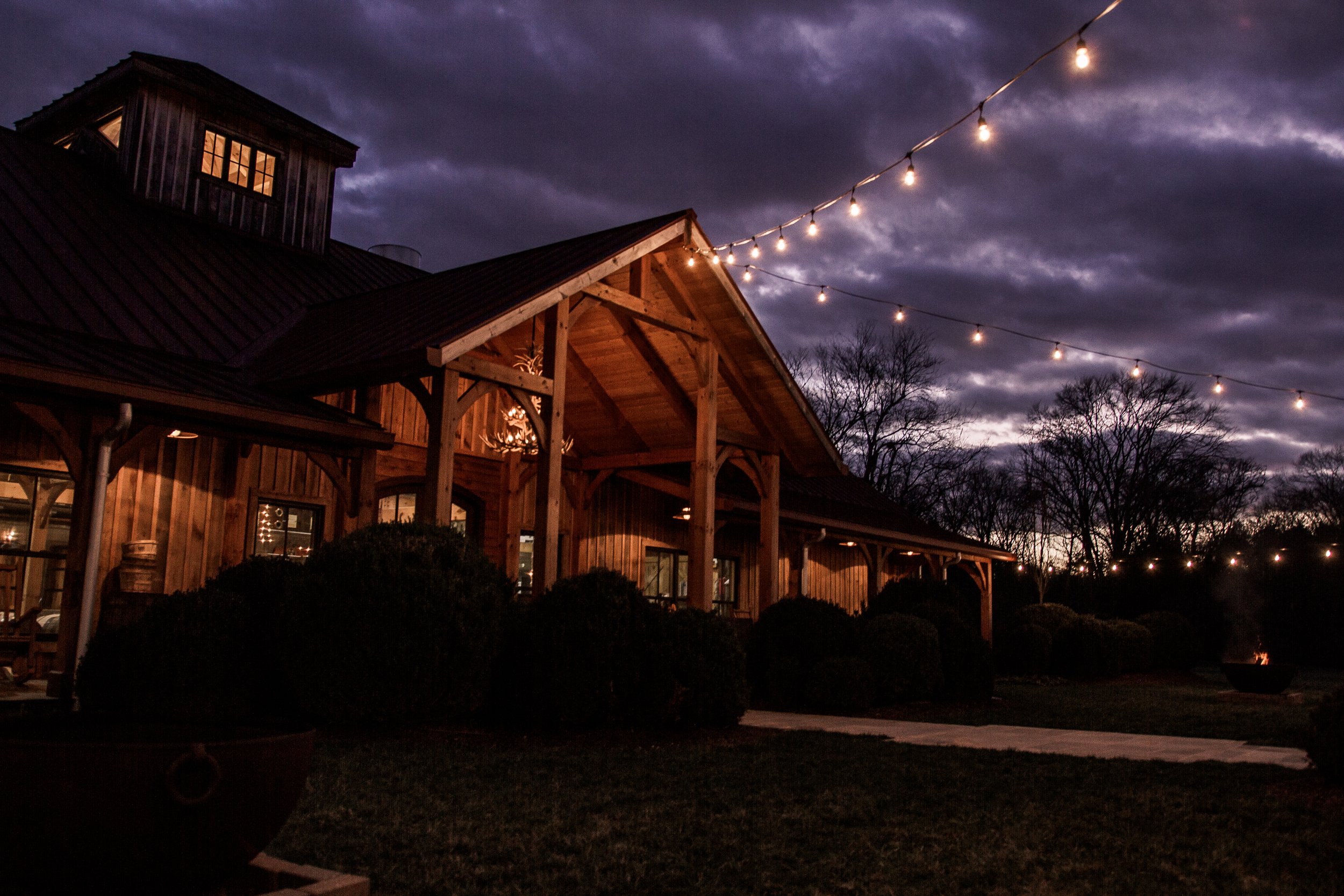 A wooden building with string lights hanging outside during dusk, with a cloudy sky and trees in the background.
