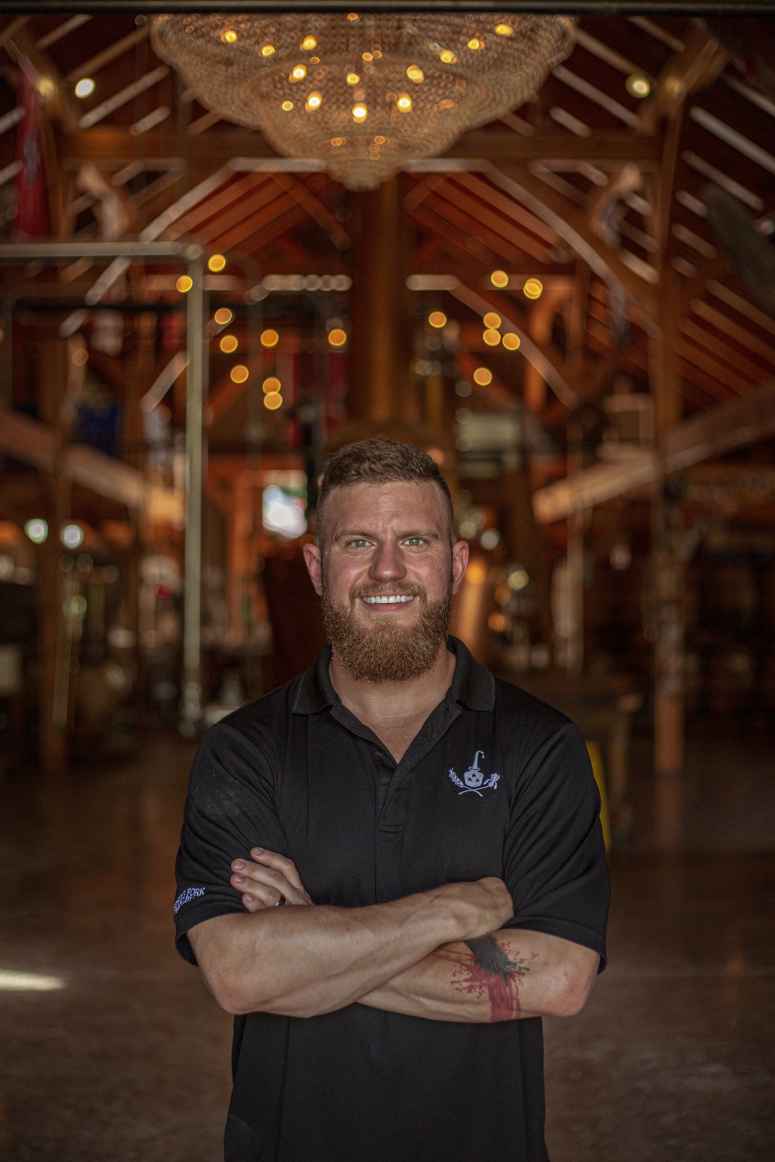 A smiling man with a beard, wearing a black polo shirt with a small emblem on the chest, standing inside a rustic wooden building with exposed beams and warm lighting.