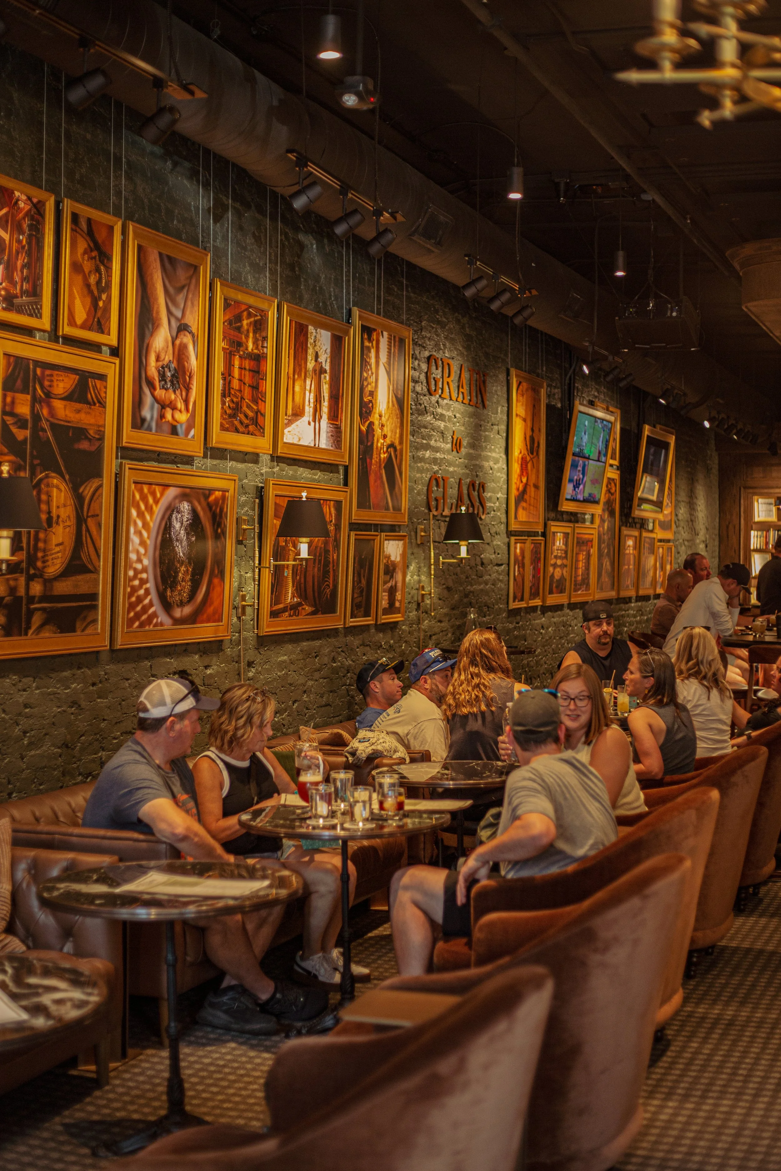 People enjoying drinks and conversation in a cozy, dimly lit restaurant or bar with brick walls decorated with framed artwork and a sign that reads 'GRAIN to GLASS'.