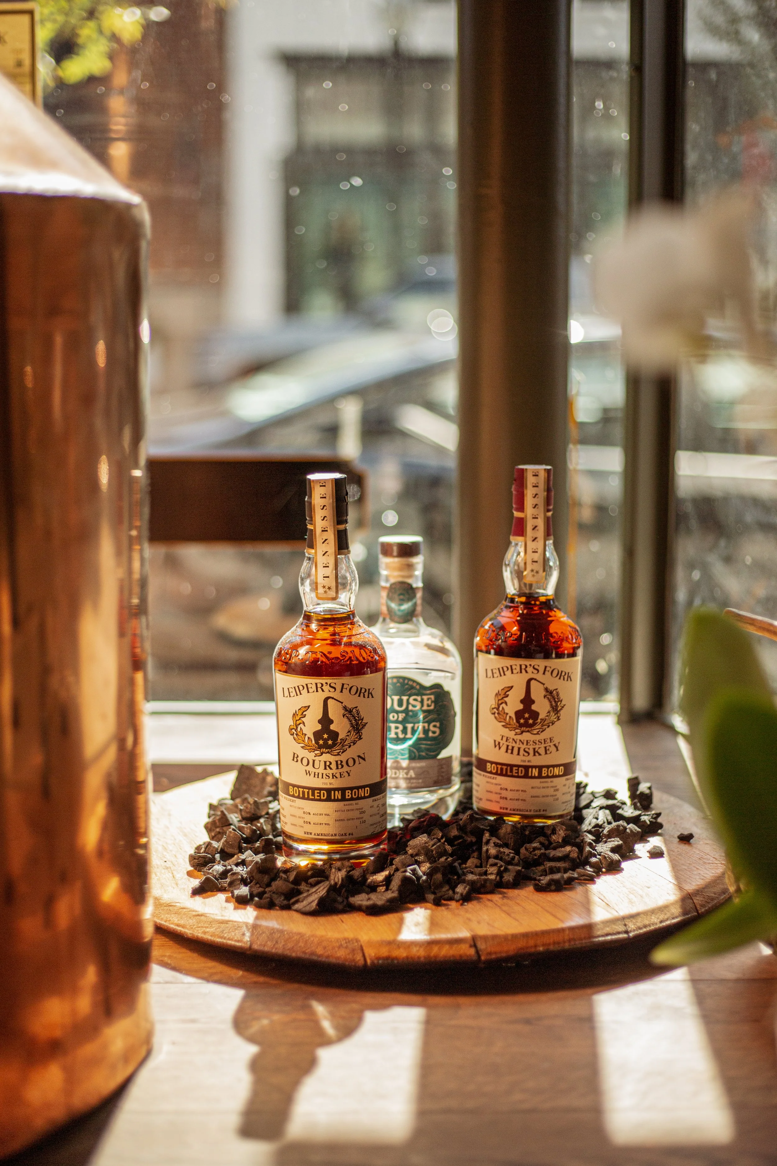 Two bottles of bourbon whiskey and a bottle of vodka on a wooden tray with black rocks, set on a table near a window.