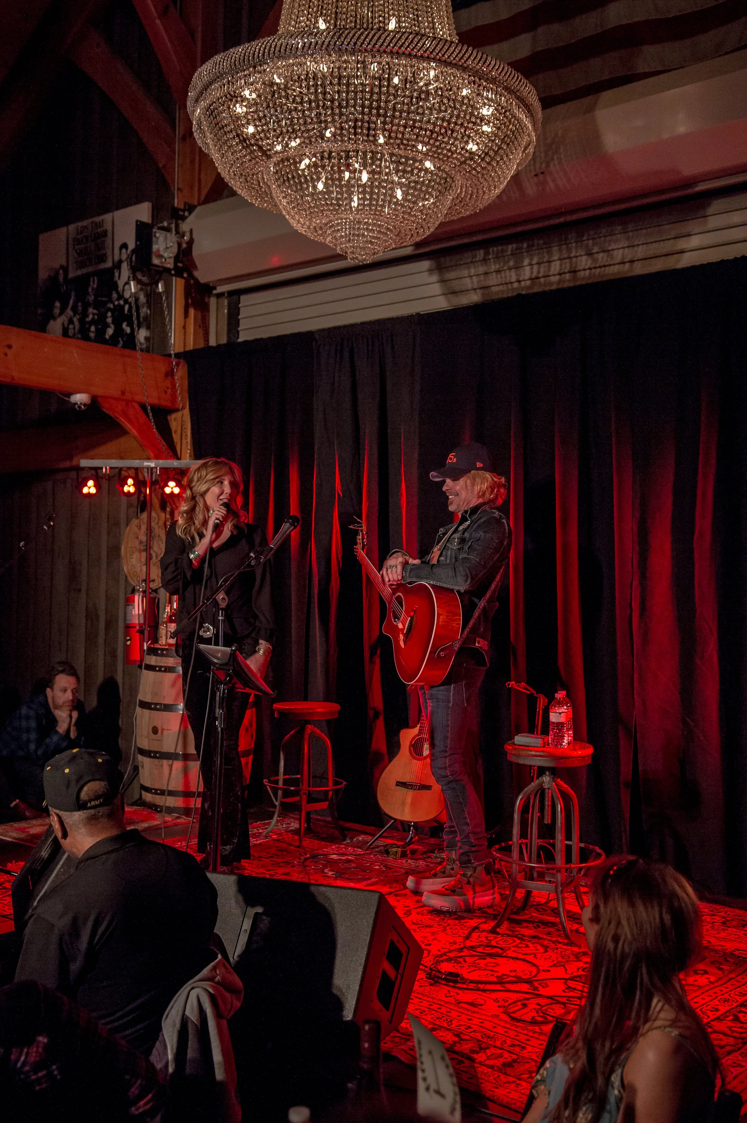 A musical performance on stage with two female performers, one singing into a microphone and the other playing an acoustic guitar, in a dimly lit venue with a chandelier overhead and an audience seated in front.