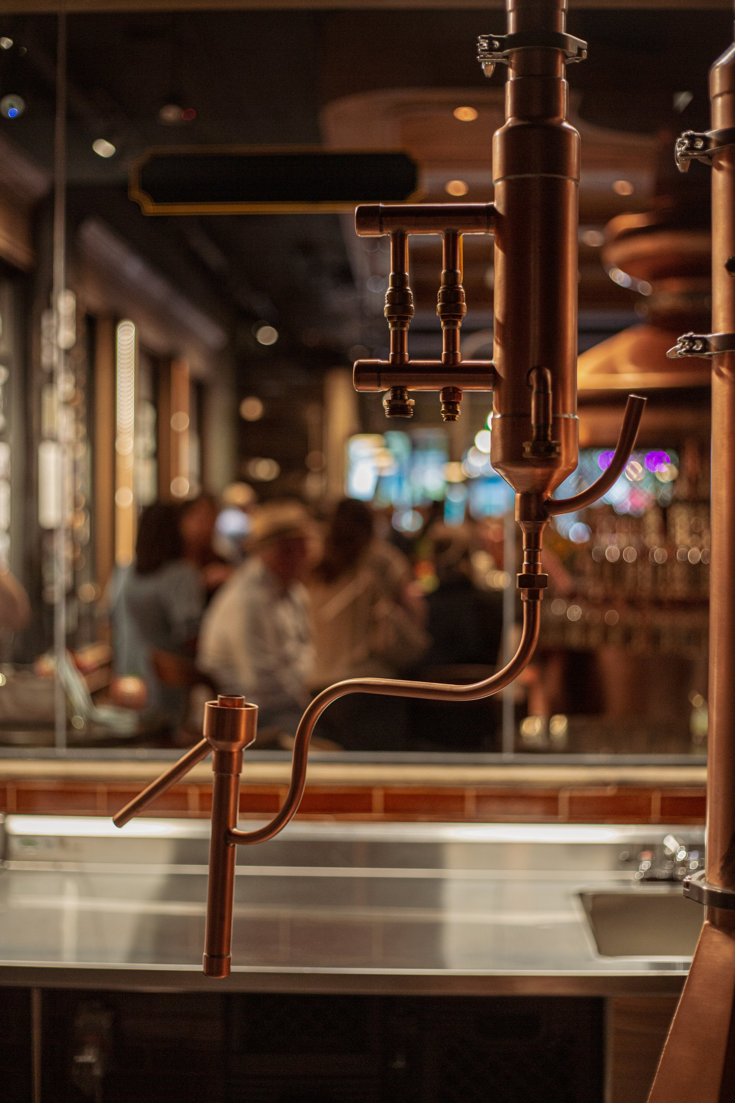 Close-up of copper brewing equipment in a bar or pub with blurred patrons in the background.