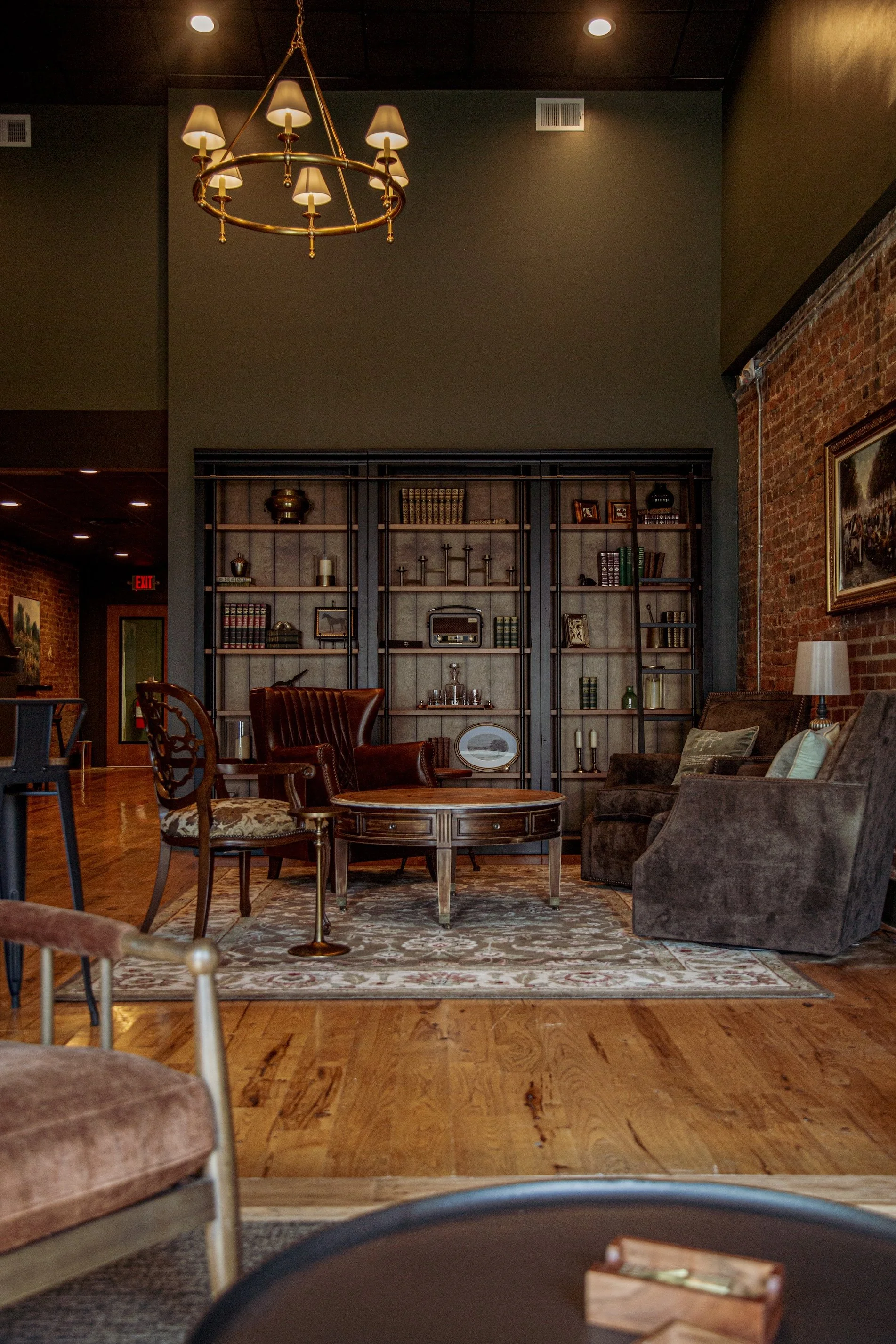 Cozy living room with vintage furniture, wooden bookshelf, and brick wall, illuminated by chandeliers and lamps.