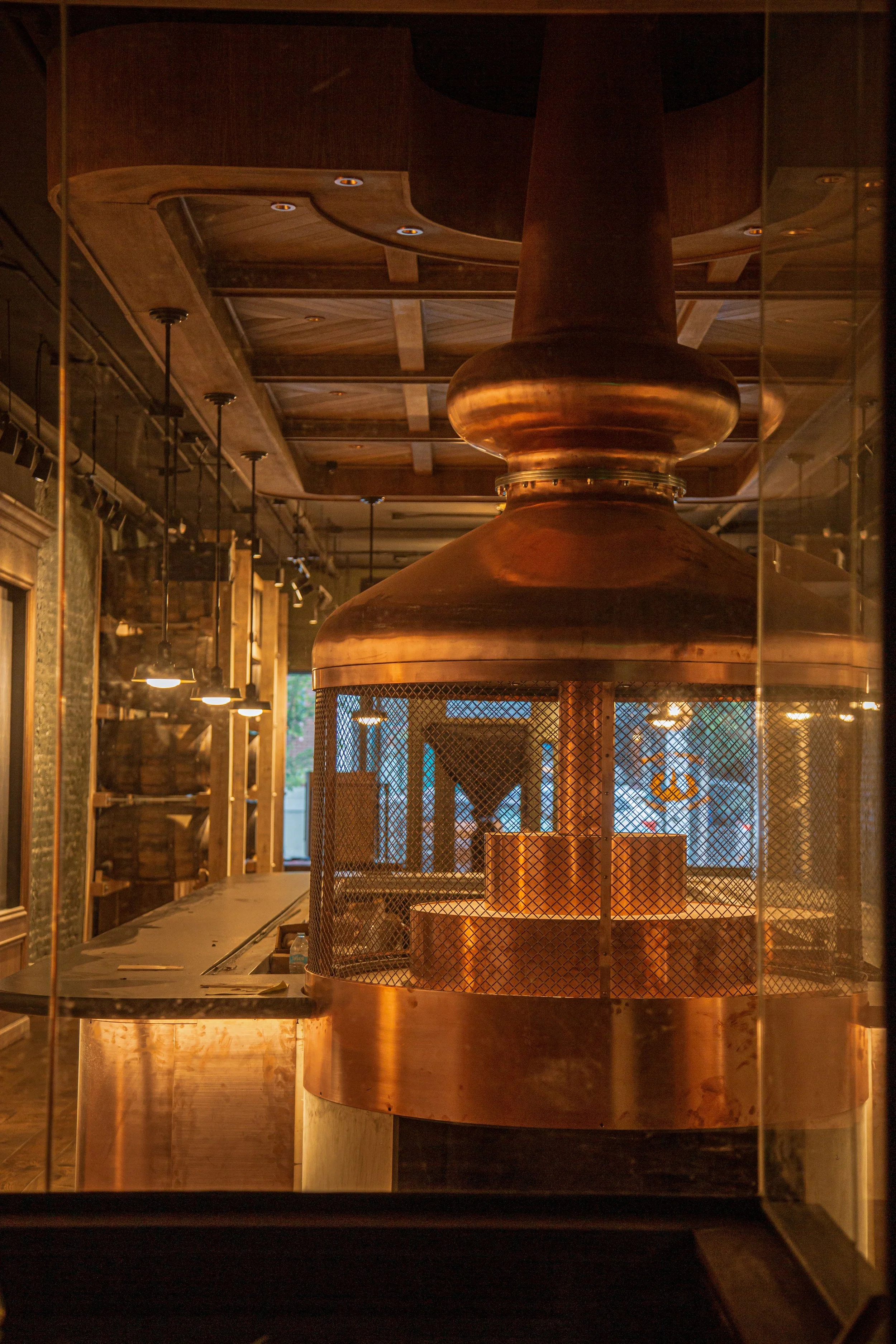 Interior of a brewery or pub featuring a large copper brewing kettle viewed through a glass window, with warm lighting and wooden decor.