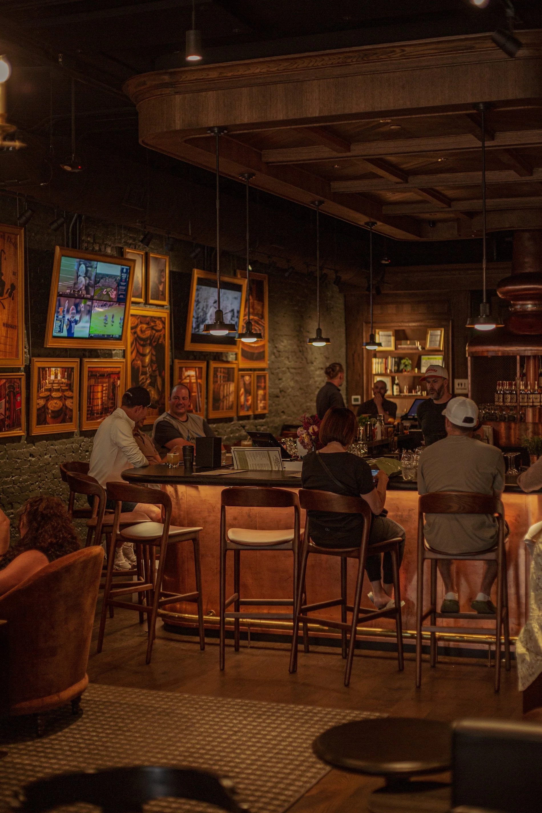 People sitting and chatting at a bar with a dark wood interior, hanging pendant lights, and framed pictures on the wall.
